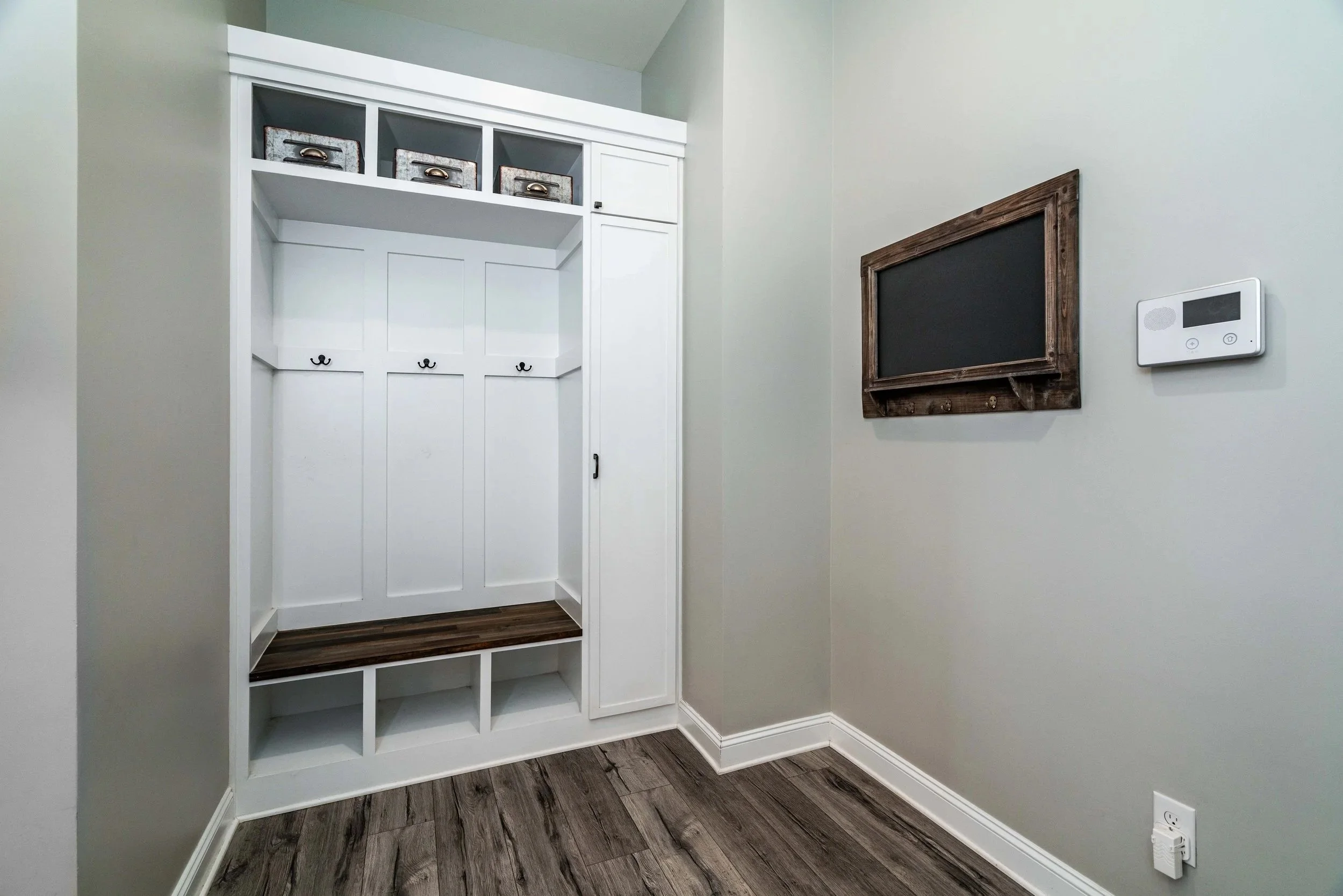 A mudroom with white built-in storage featuring three hooks and a wooden bench. Shelves above hold baskets. A chalkboard and thermostat on a gray wall.