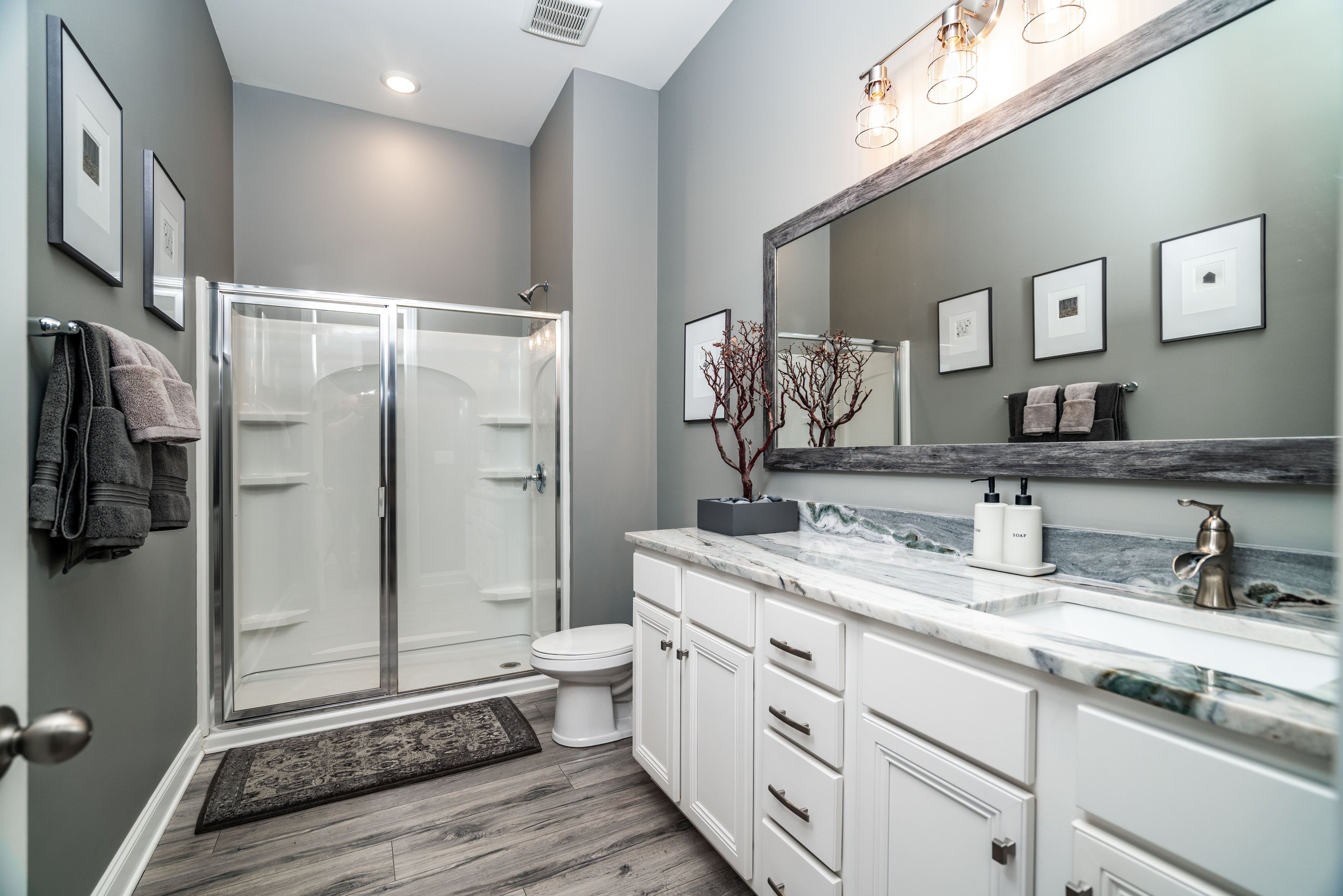 Modern bathroom with gray walls, glass shower, and dual sinks on a marble countertop. Decor includes monochrome art, towels, and a plant, creating a serene feel.
