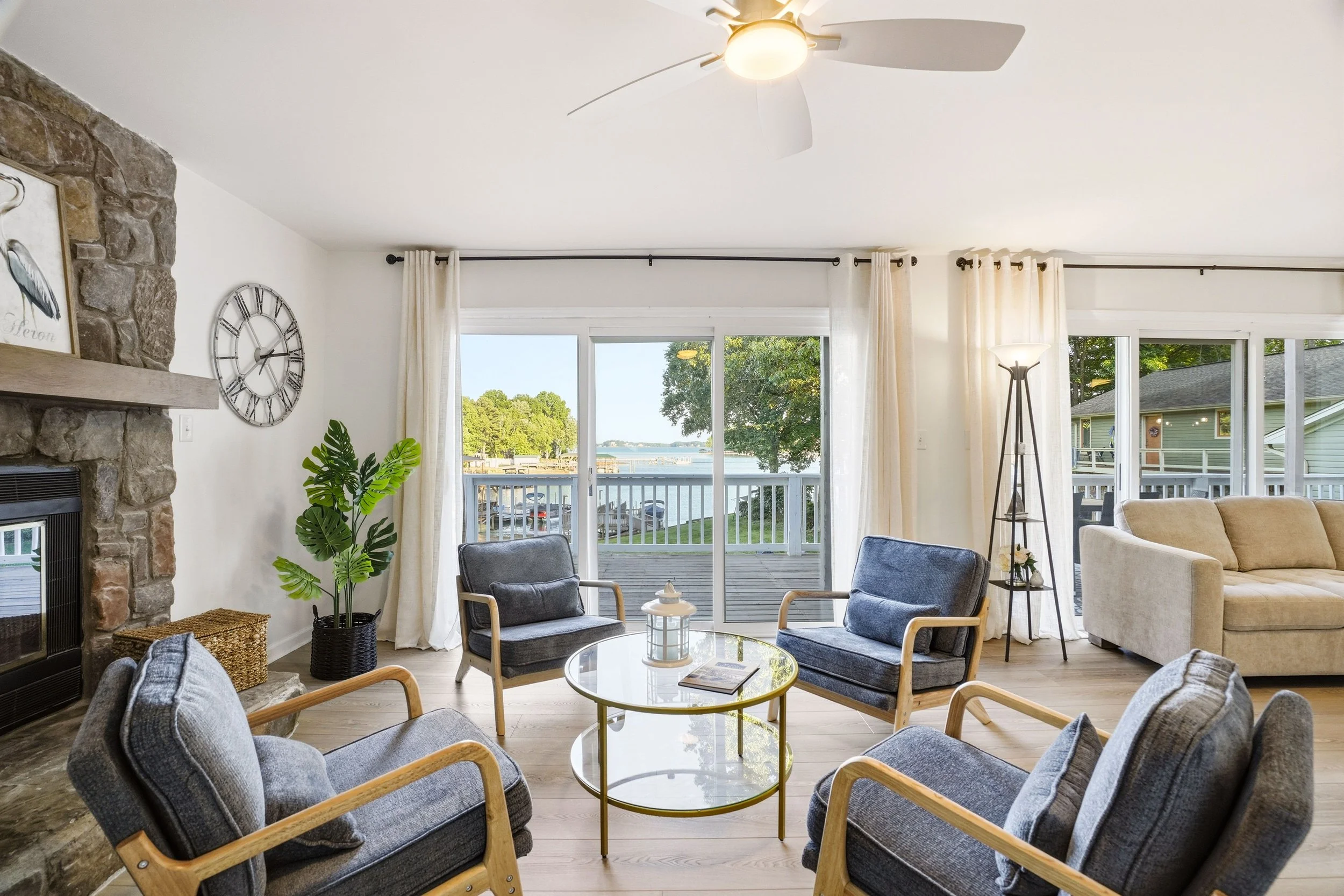 A modern living room with four grey armchairs around a glass coffee table, facing a large stone fireplace. Sliding glass doors reveal a lake with boat docks outside.