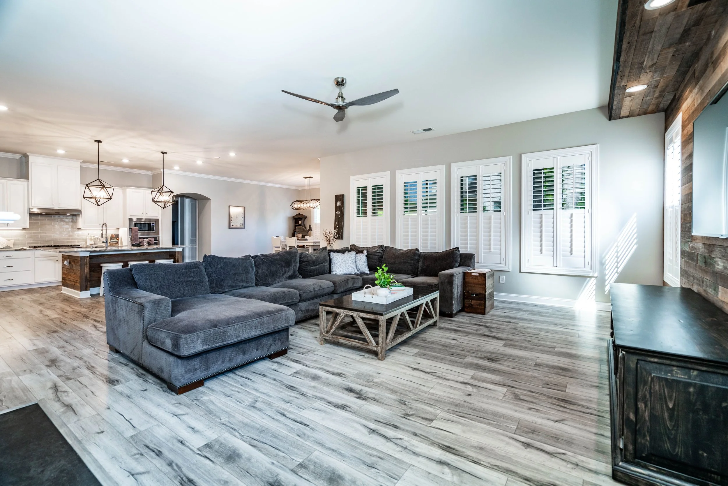 Modern living room with gray sectional sofa facing a rustic wood coffee table. Open kitchen with pendant lights in the background. Bright, airy ambience.