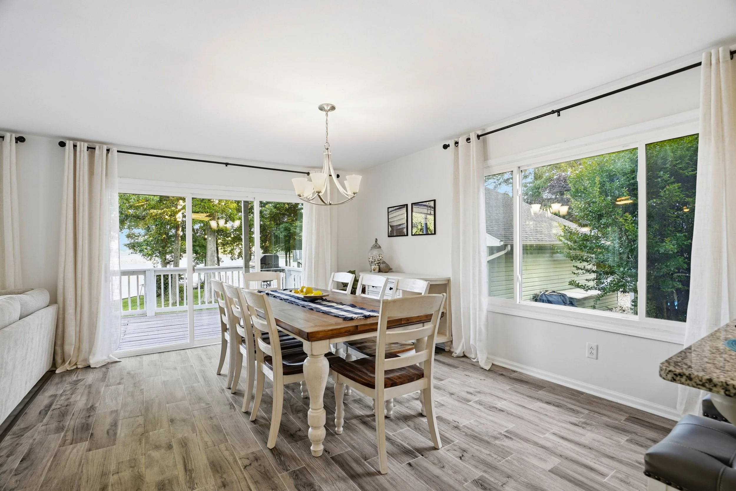 A bright dining area featuring a large wooden table surrounded by eight chairs. Natural light floods in through large windows, with white curtains hanging beside them. The room has light wood flooring and a chandelier above the table.