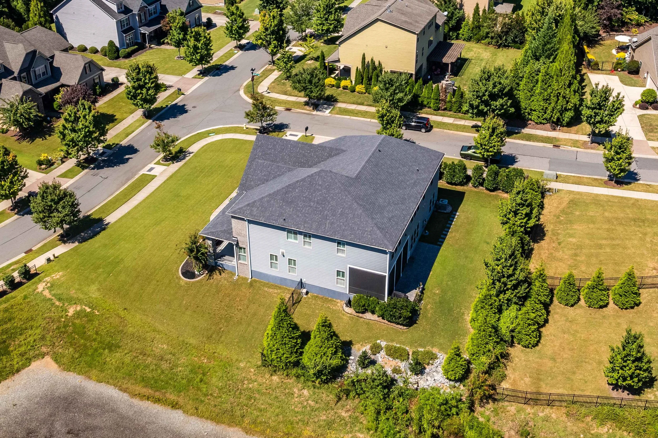 Aerial view of a suburban neighborhood featuring a large house with a dark roof centered on a spacious green lawn. Tree-lined streets and houses surround it.