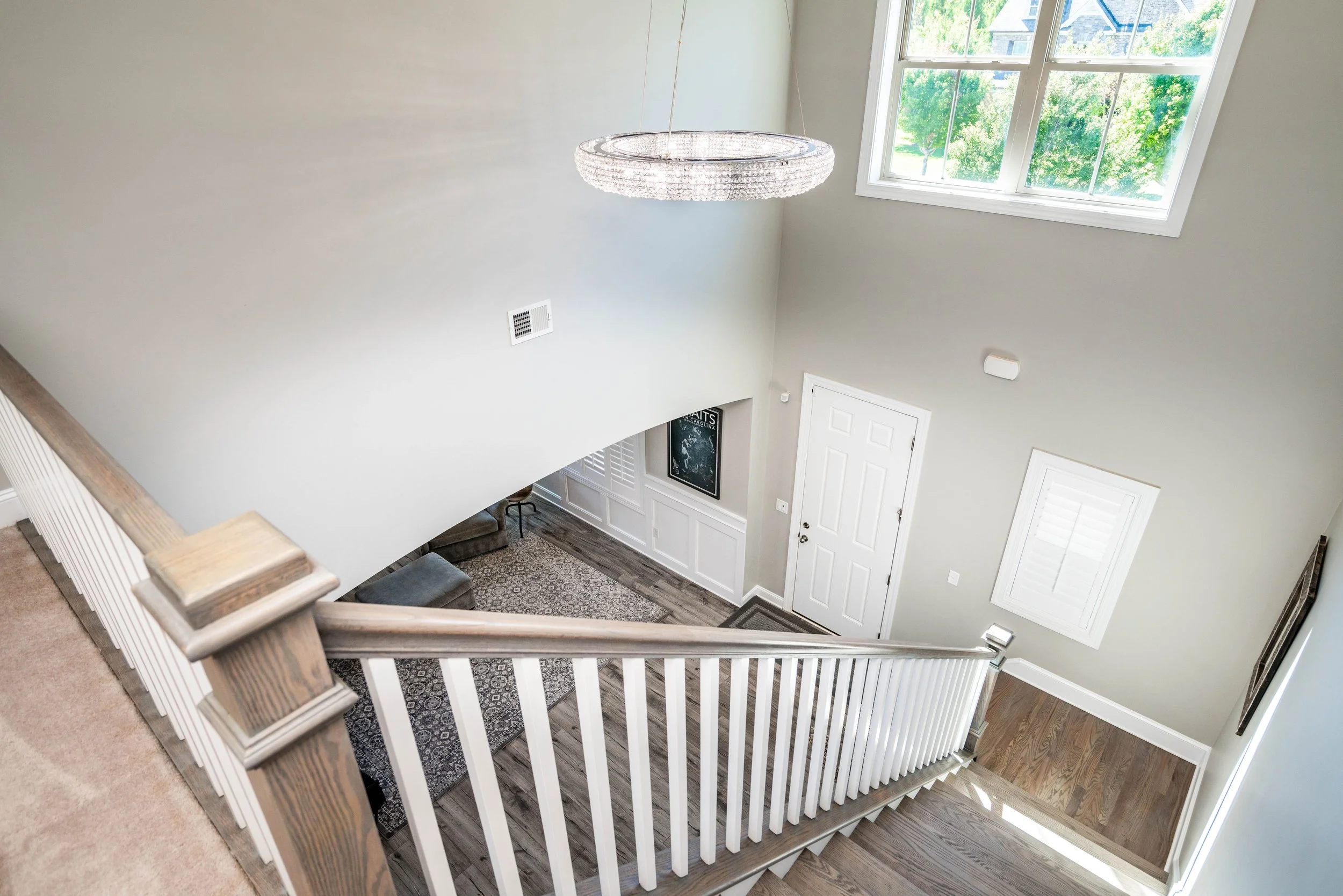 A modern staircase with white railings leads down to an elegant foyer. A large window brightens the space, highlighting a sleek chandelier and white door.