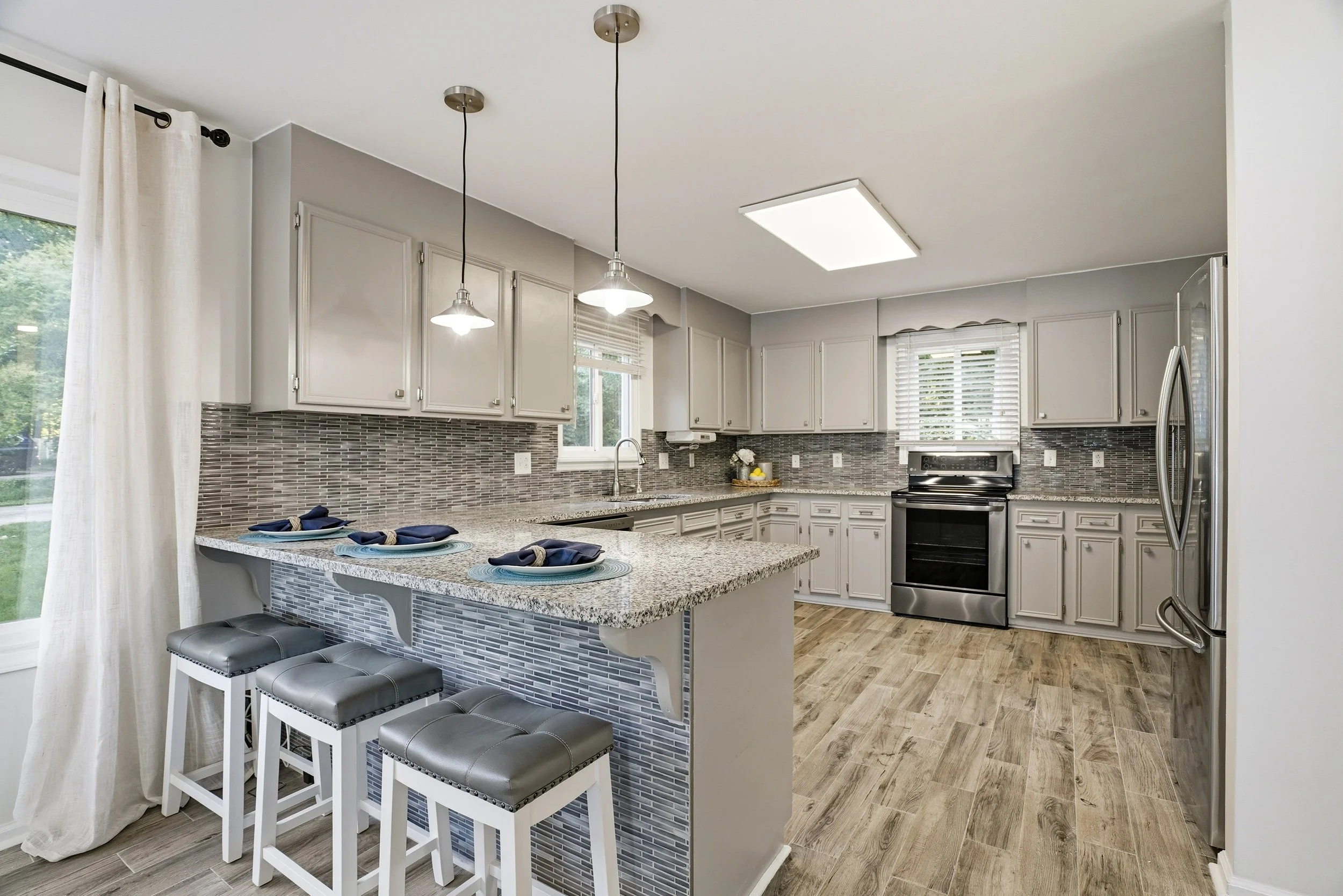A modern kitchen with light gray cabinets and a gray and white tiled backsplash. A central island with a speckled countertop features three white barstools, and stainless steel appliances are visible, including an oven and refrigerator.