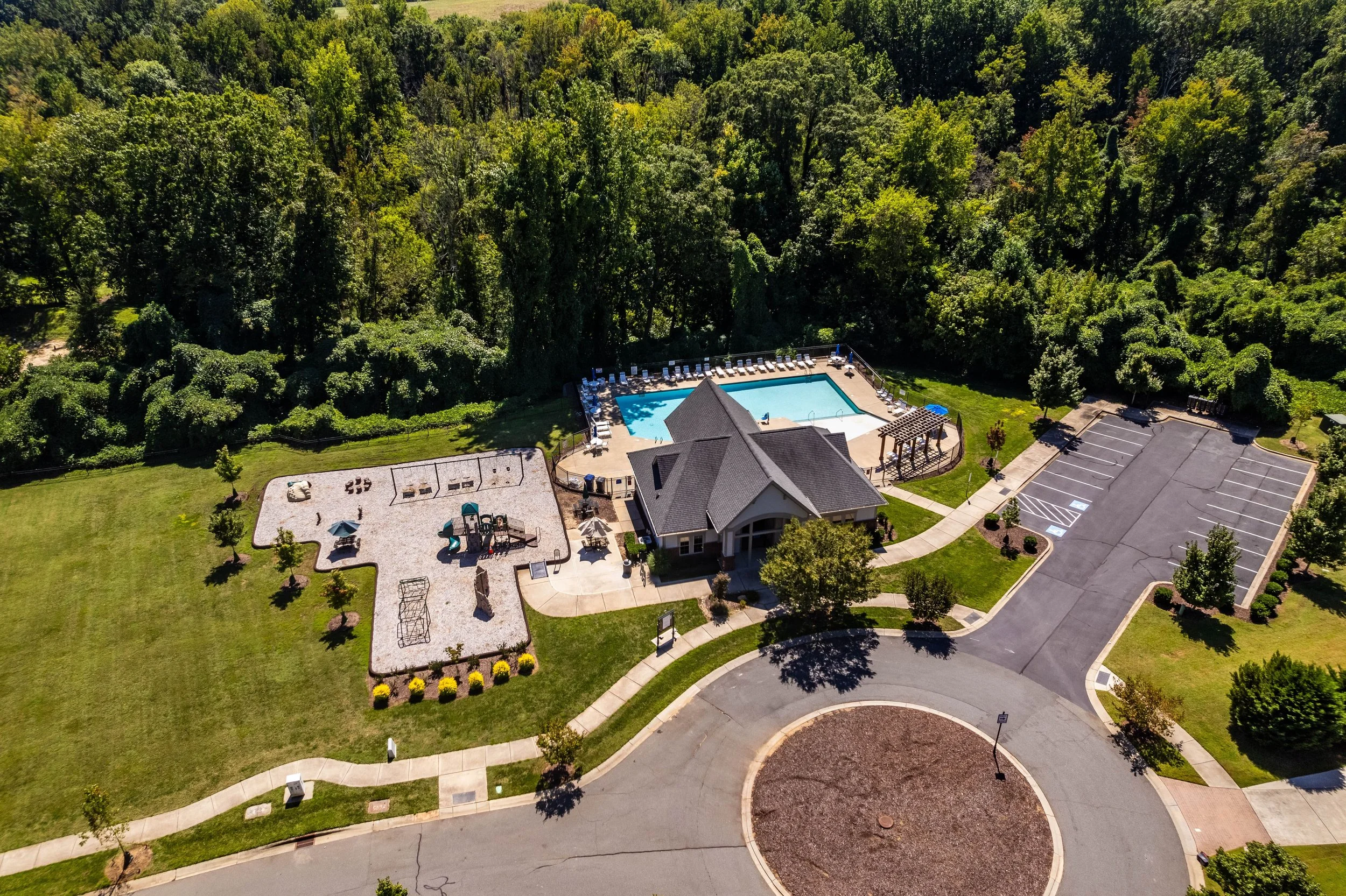 Aerial view of a community recreation area. Features a swimming pool, playground, parking lot, and clubhouse, surrounded by lush green trees and grass.