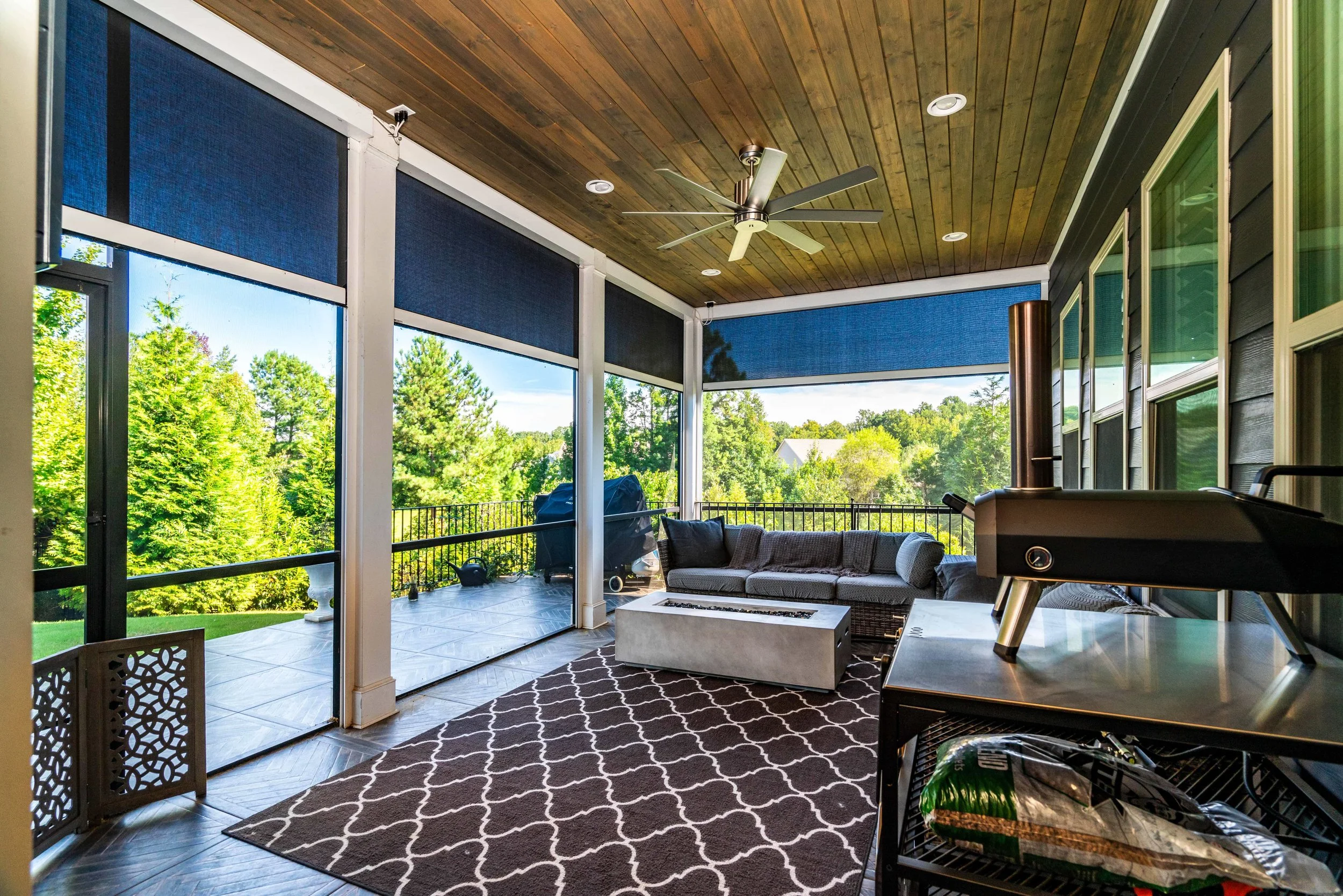 Covered patio with a wooden ceiling, ceiling fan, and large windows overlooking greenery. Features a cushioned sofa, patterned rug, and outdoor grill.