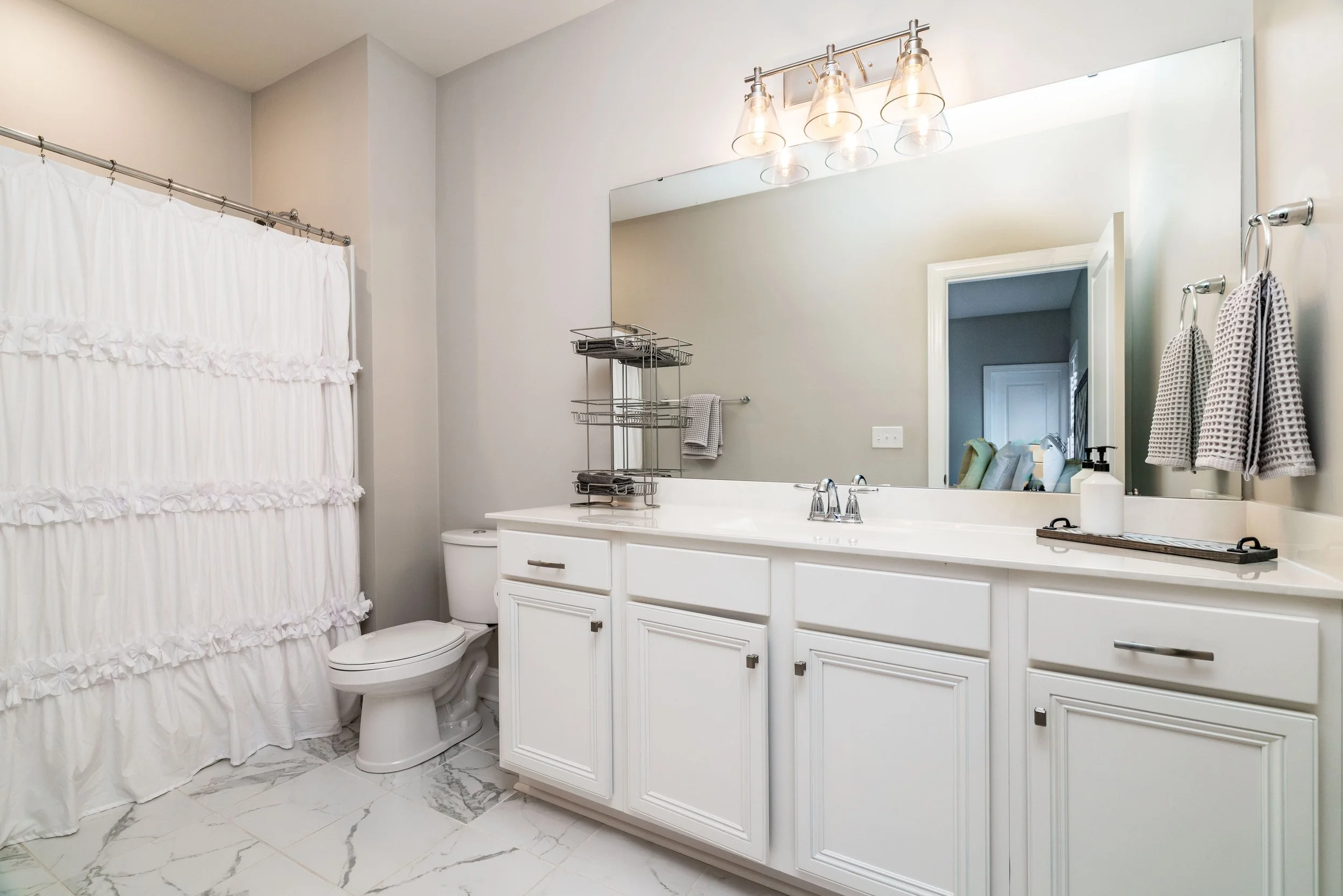 Bright, modern bathroom with white cabinetry and marble flooring. A large mirror and three-light fixture above the sink enhance the clean, fresh ambiance.