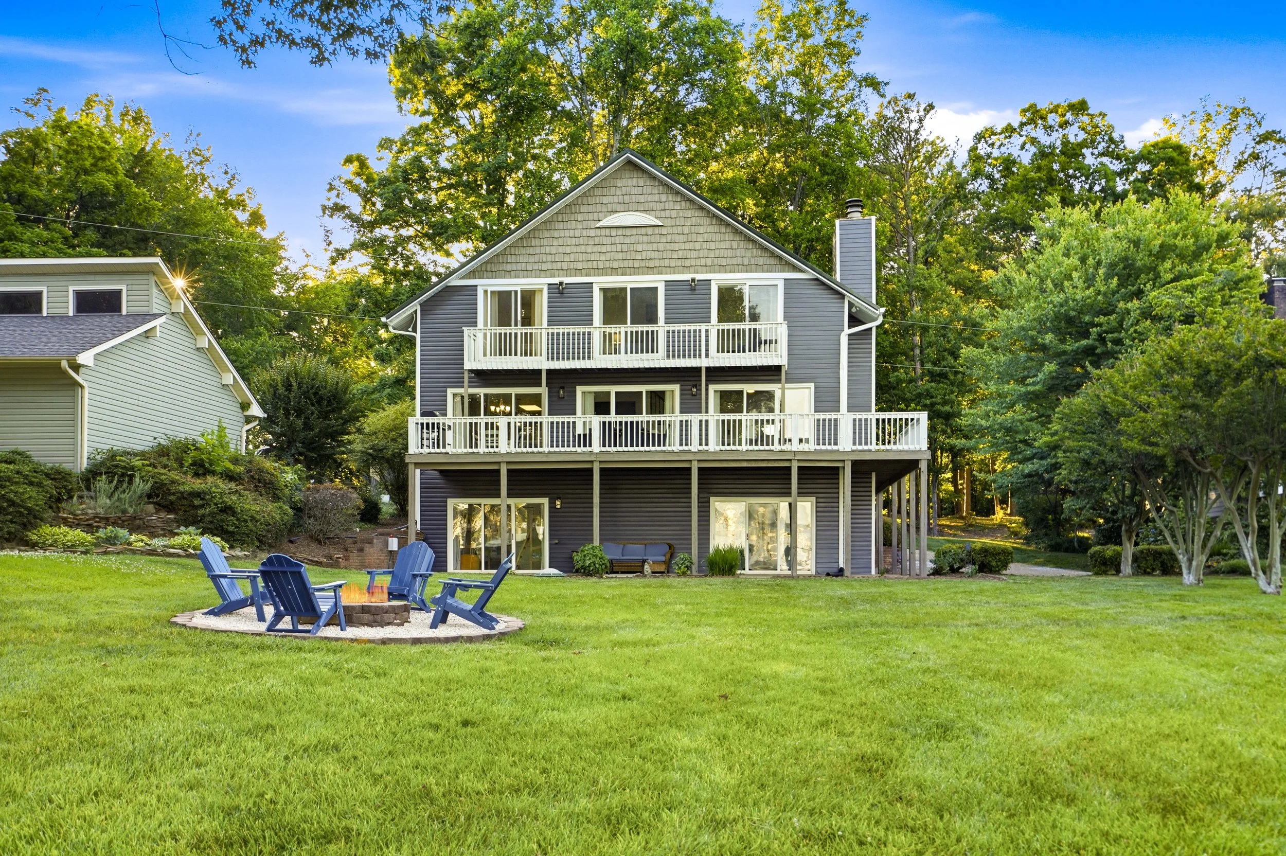 Two-story house with large balcony and patio, surrounding lawn, trees, and a circular fire pit area with blue chairs in the backyard under a clear sky.