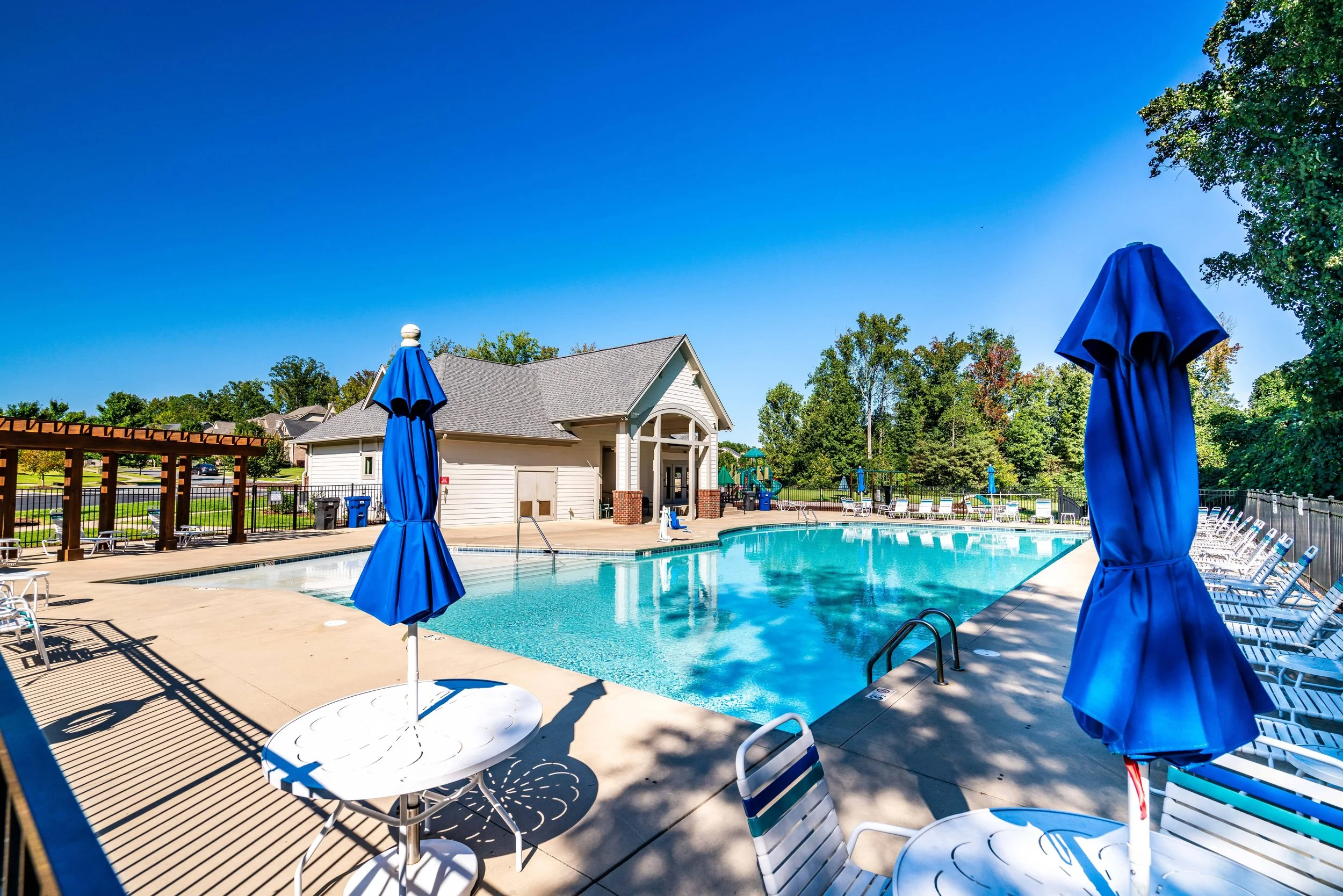 A serene outdoor pool under a clear blue sky, flanked by white lounge chairs and tables with closed blue umbrellas. A cozy clubhouse in the background.