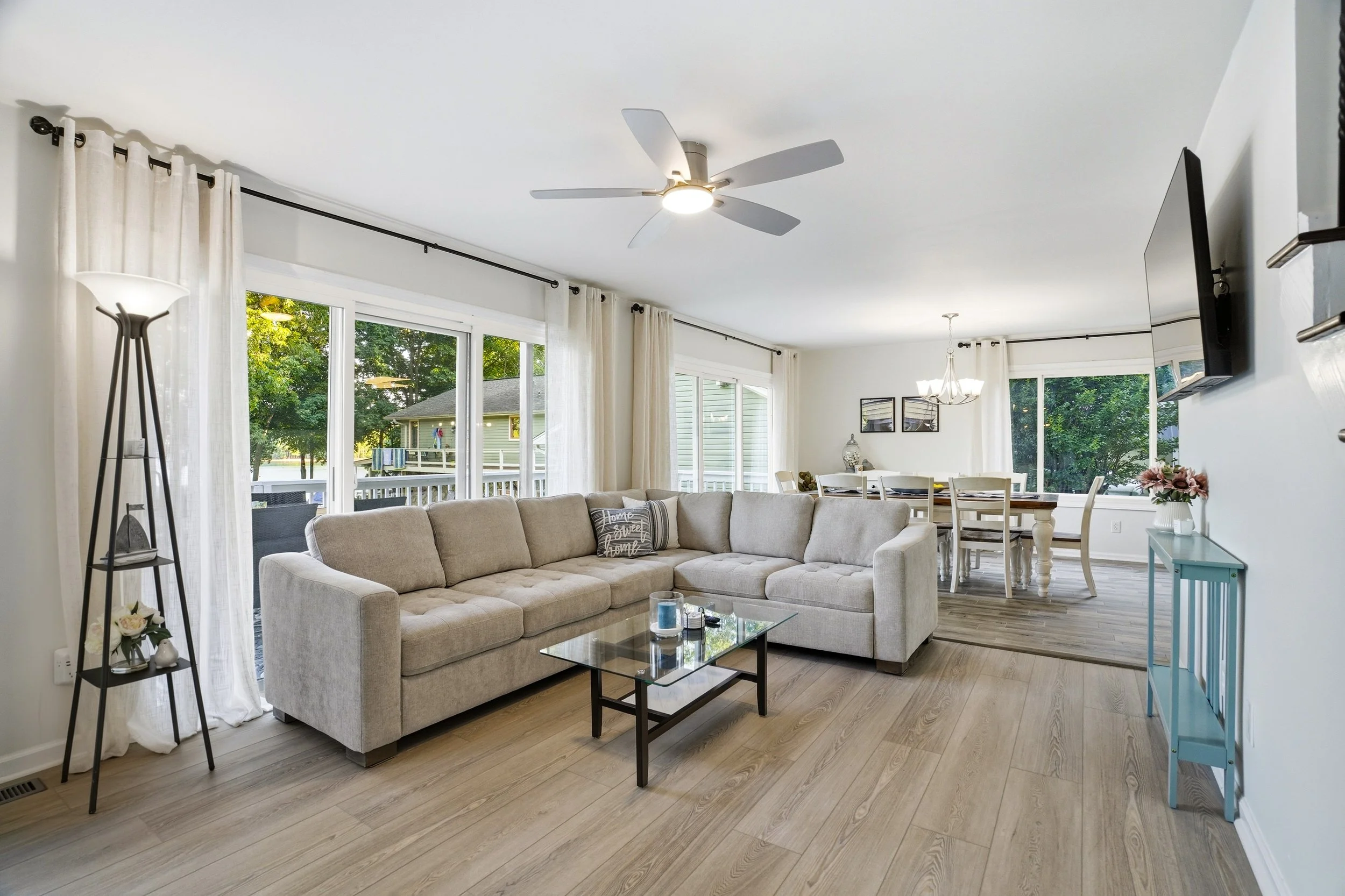 A modern living room with a light gray sectional sofa and a glass coffee table. Two large windows with white curtains let in natural light, and a dining area with a table and chairs is visible in the background. A ceiling fan is above.