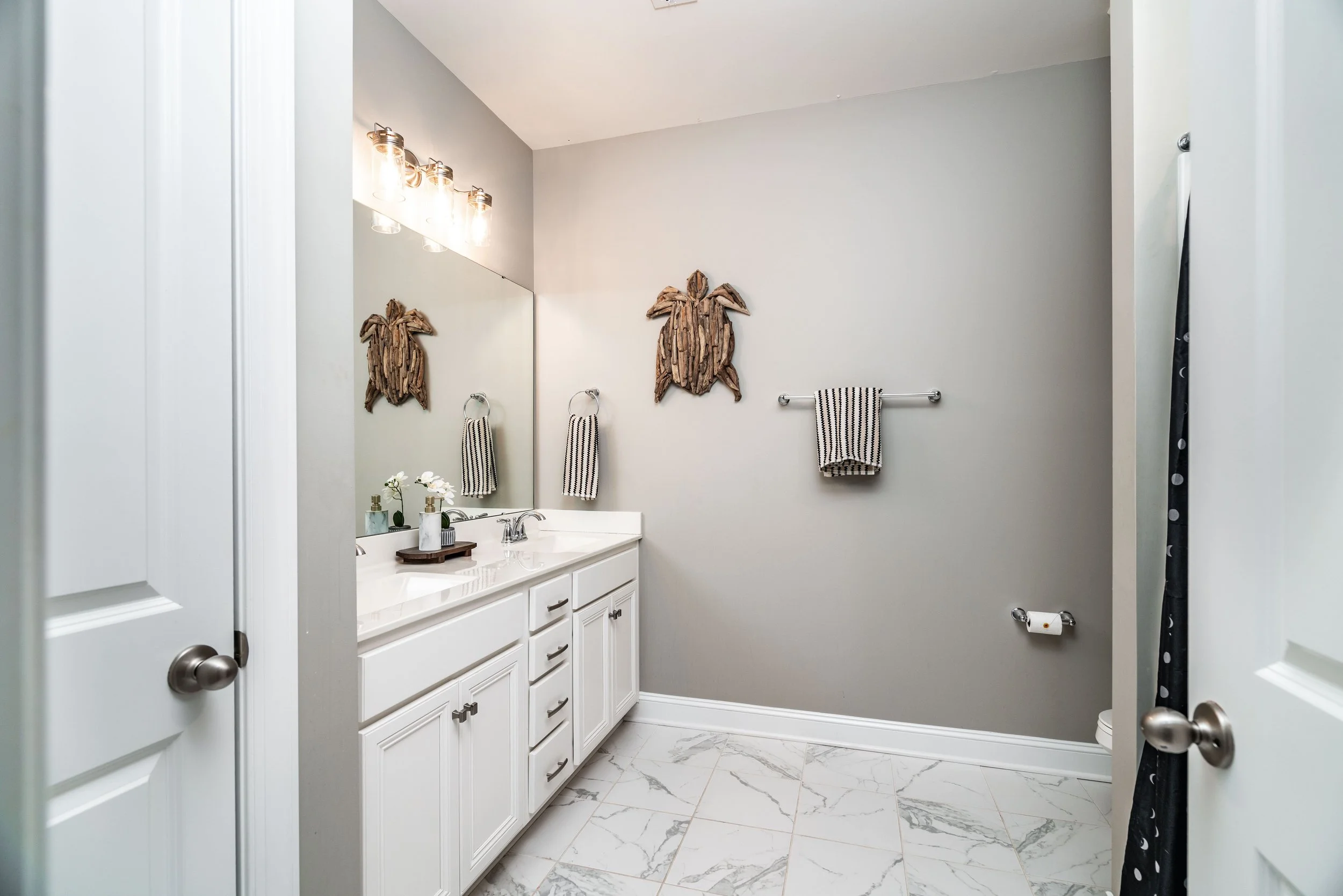 A modern bathroom with white cabinetry, a marble countertop, and polished fixtures. A turtle sculpture and striped towels add decorative touches on gray walls.