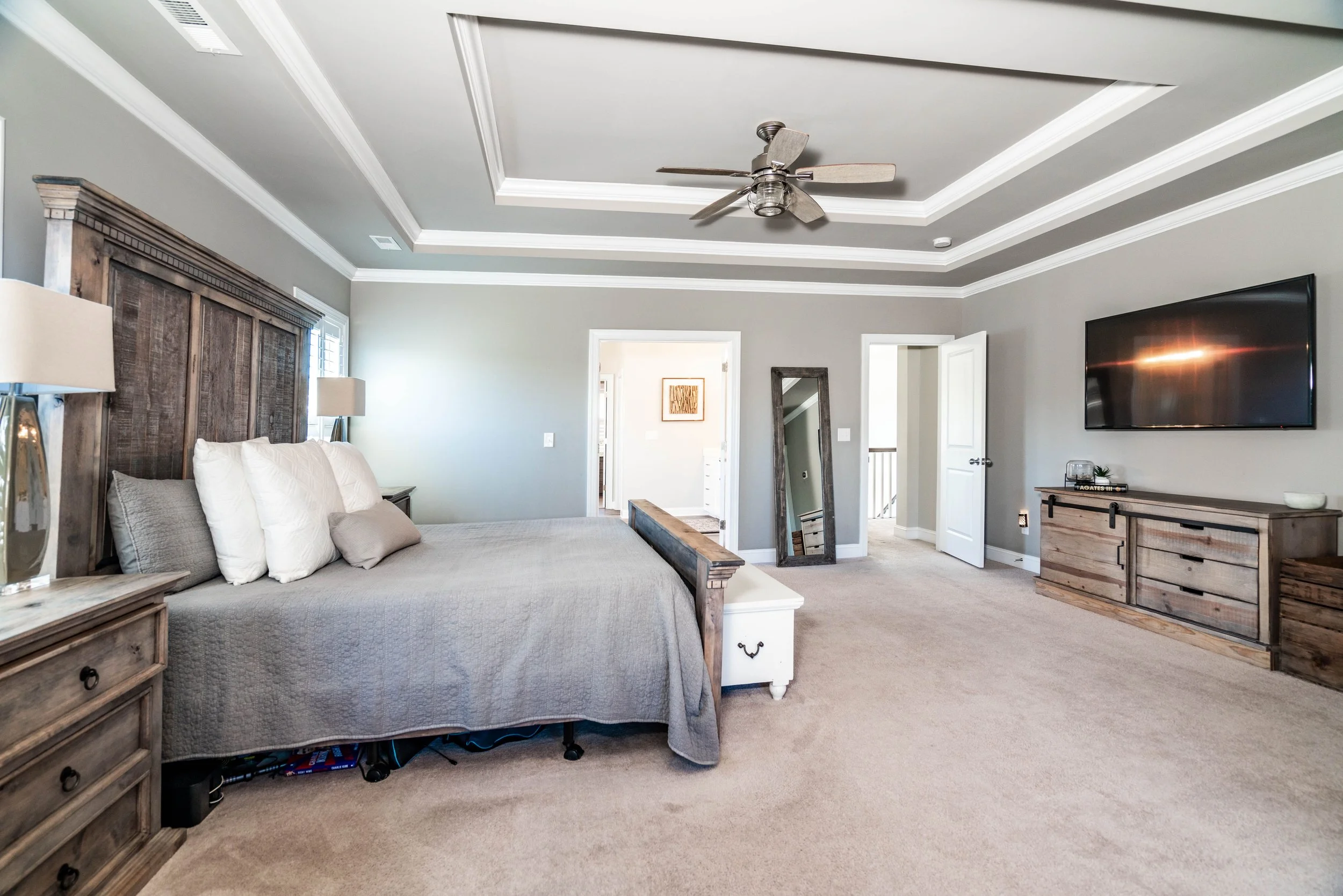 Spacious bedroom with a gray color scheme, featuring a large bed with white pillows, wooden furniture, a ceiling fan, and a mounted TV, conveying comfort.