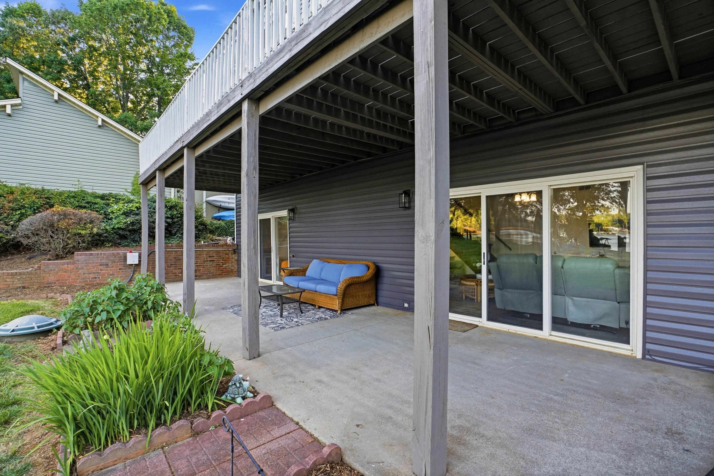Covered concrete patio beneath a raised deck, featuring a blue wicker couch and sliding glass doors leading indoors against dark gray siding.
