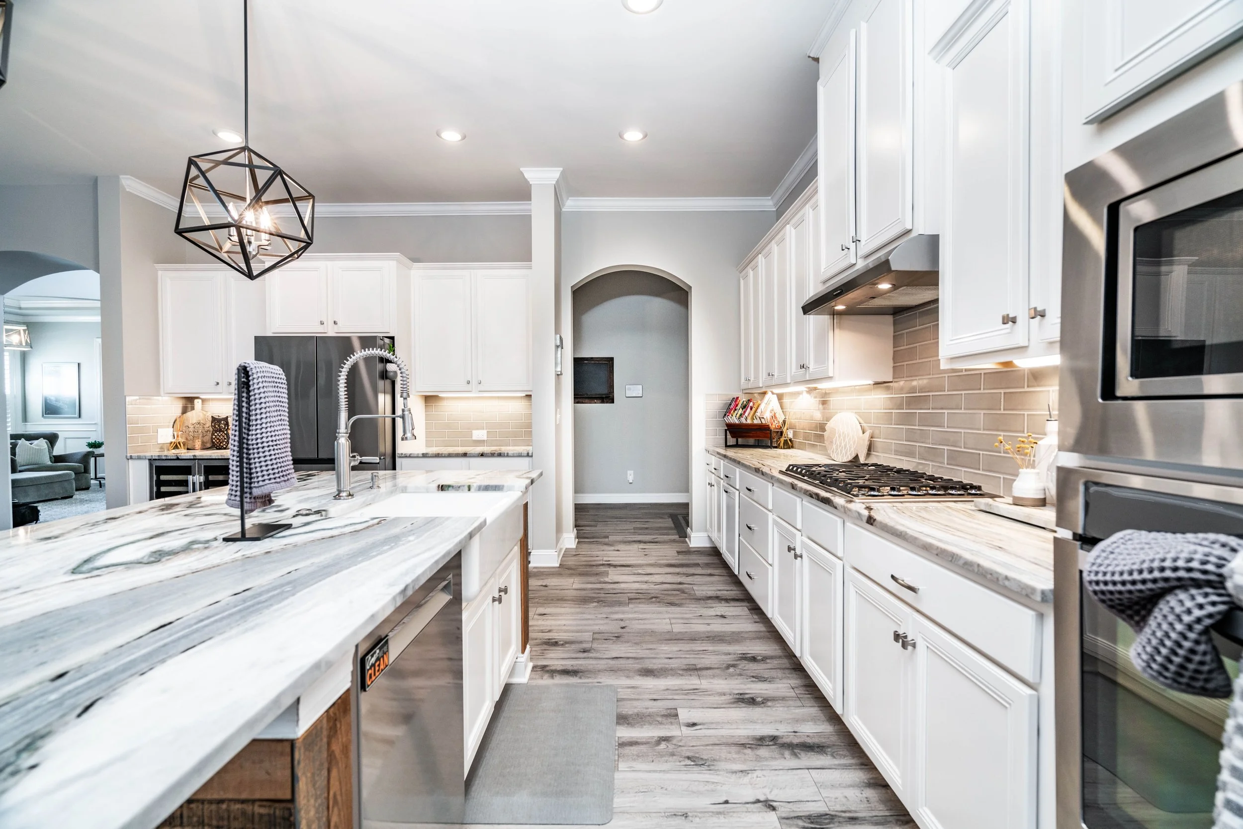 A modern kitchen with white cabinets, gray countertops, and a stainless steel oven. A geometric light fixture hangs above a spacious island, conveying a sleek, bright atmosphere.