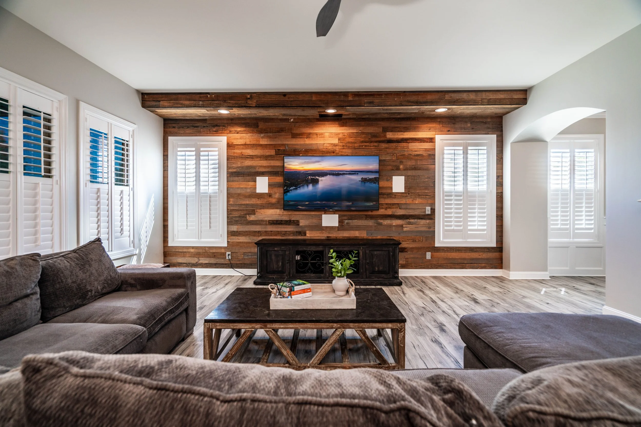Cozy living room with wood accent wall, flat-screen TV, gray sofas, rustic coffee table, and white shutters on windows, conveying a warm, inviting atmosphere.