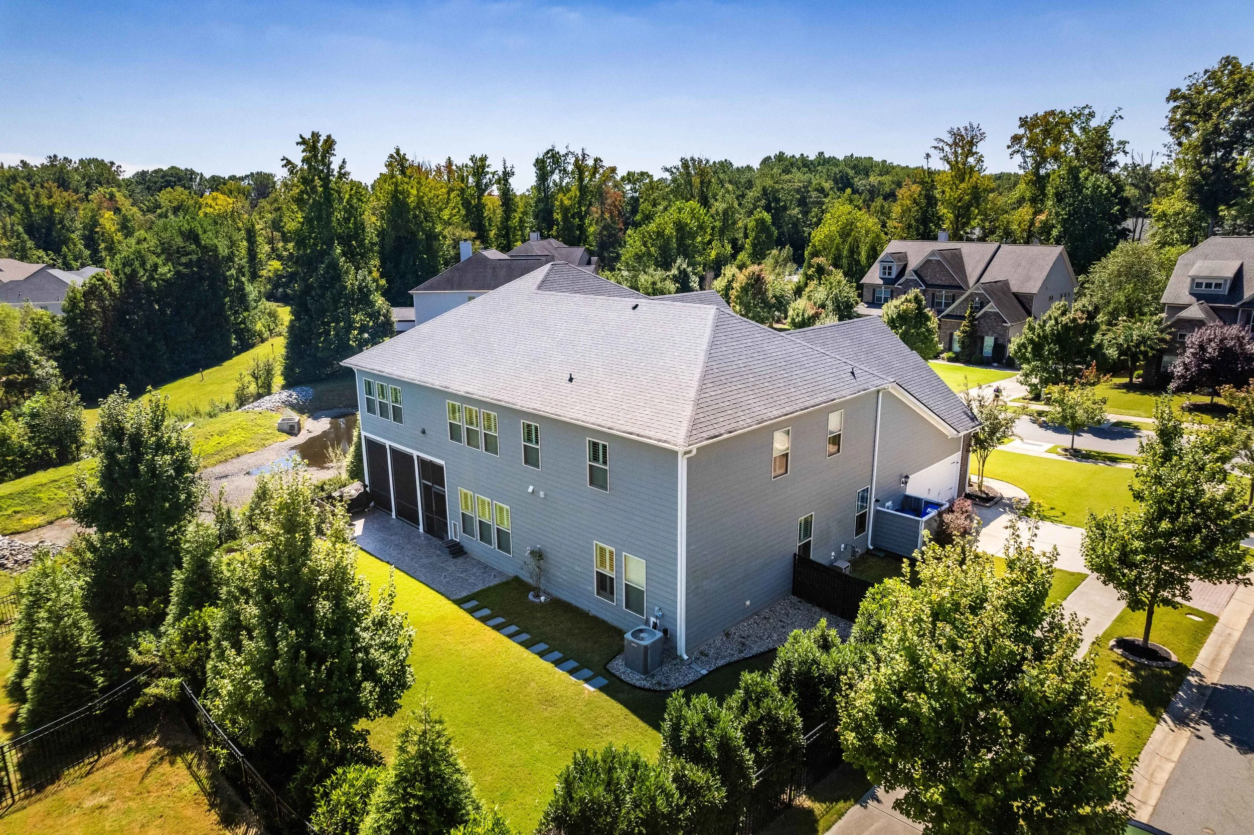 Aerial view of a large, light gray two-story house with a sloped roof, surrounded by green lawns, trees, and neighboring houses on a sunny day.