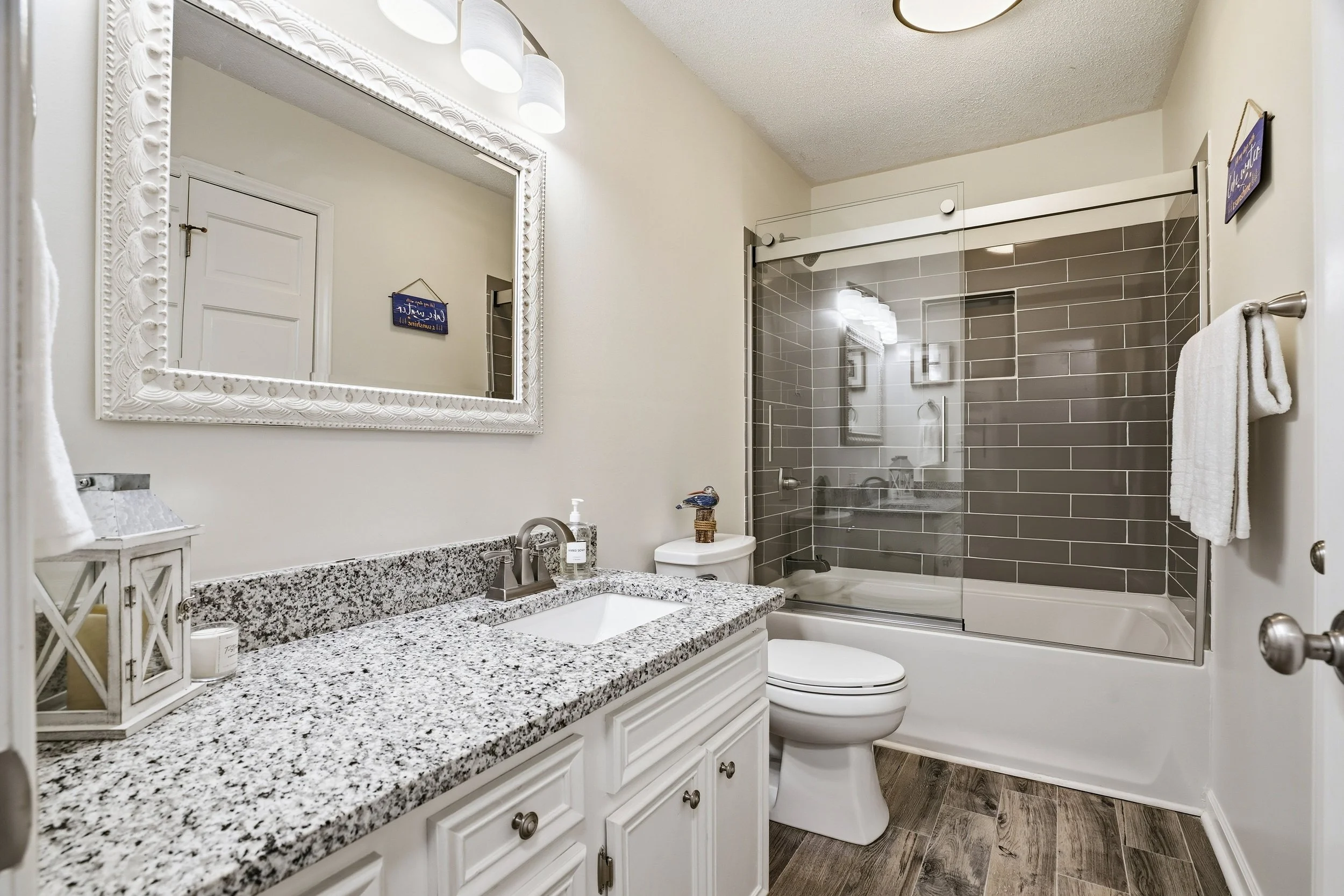 A brightly lit modern bathroom features a white vanity with granite countertops, a framed mirror, a toilet, and a tiled shower-tub combination.