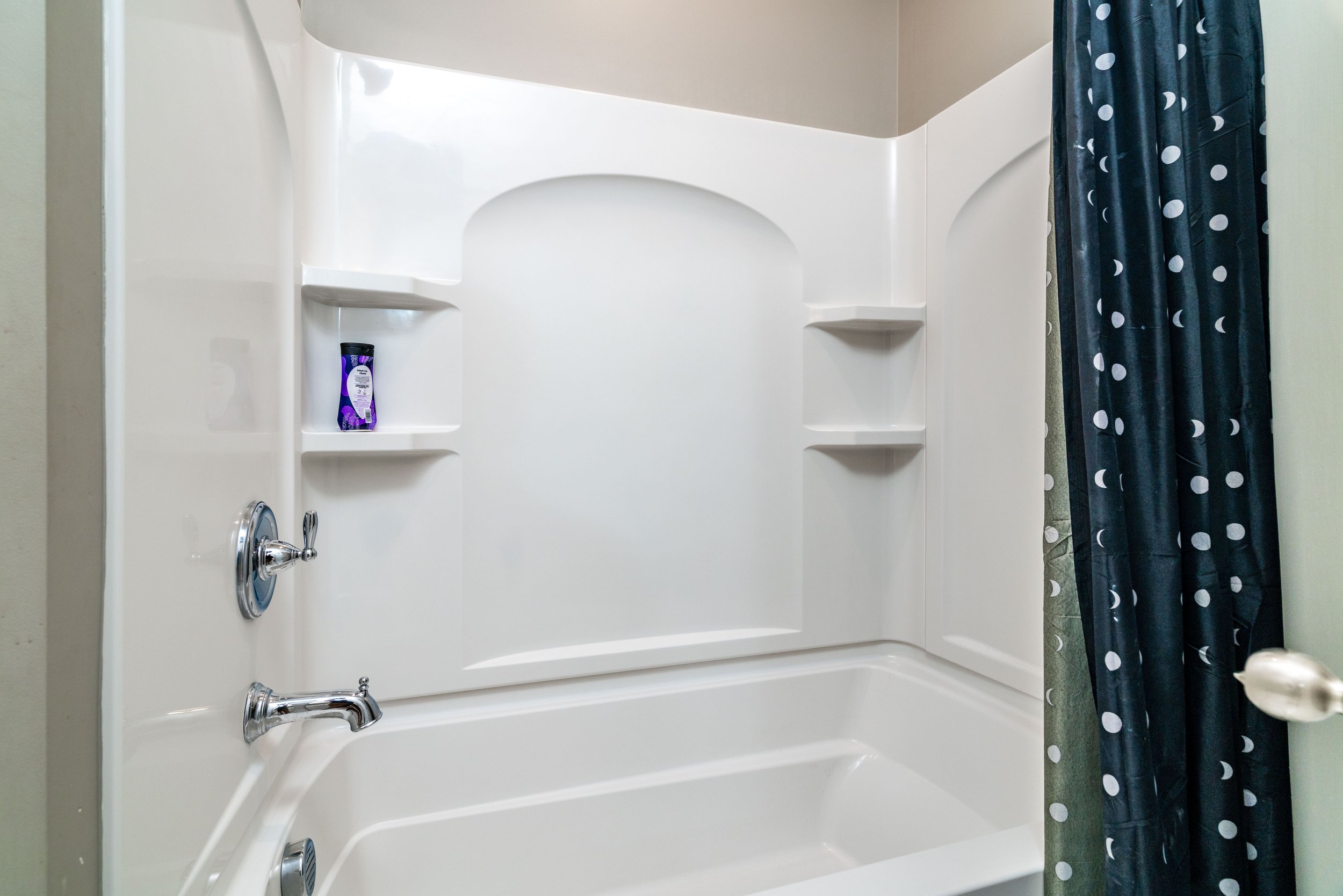 White bathroom with a bathtub, chrome fixtures, and a polka dot shower curtain. A shampoo bottle sits on a shelf, conveying a clean, minimalist atmosphere.