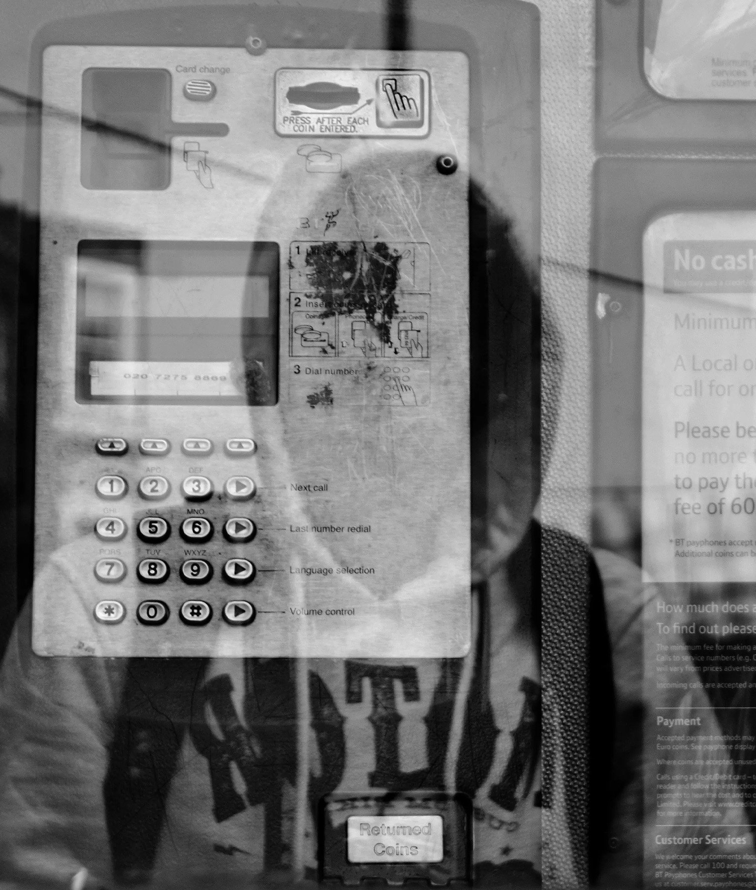 Schwarzweiss Bild: Junger Mann mit Kapuzenpullover spiegelt sich in Scheibe einer Telefonkabine
