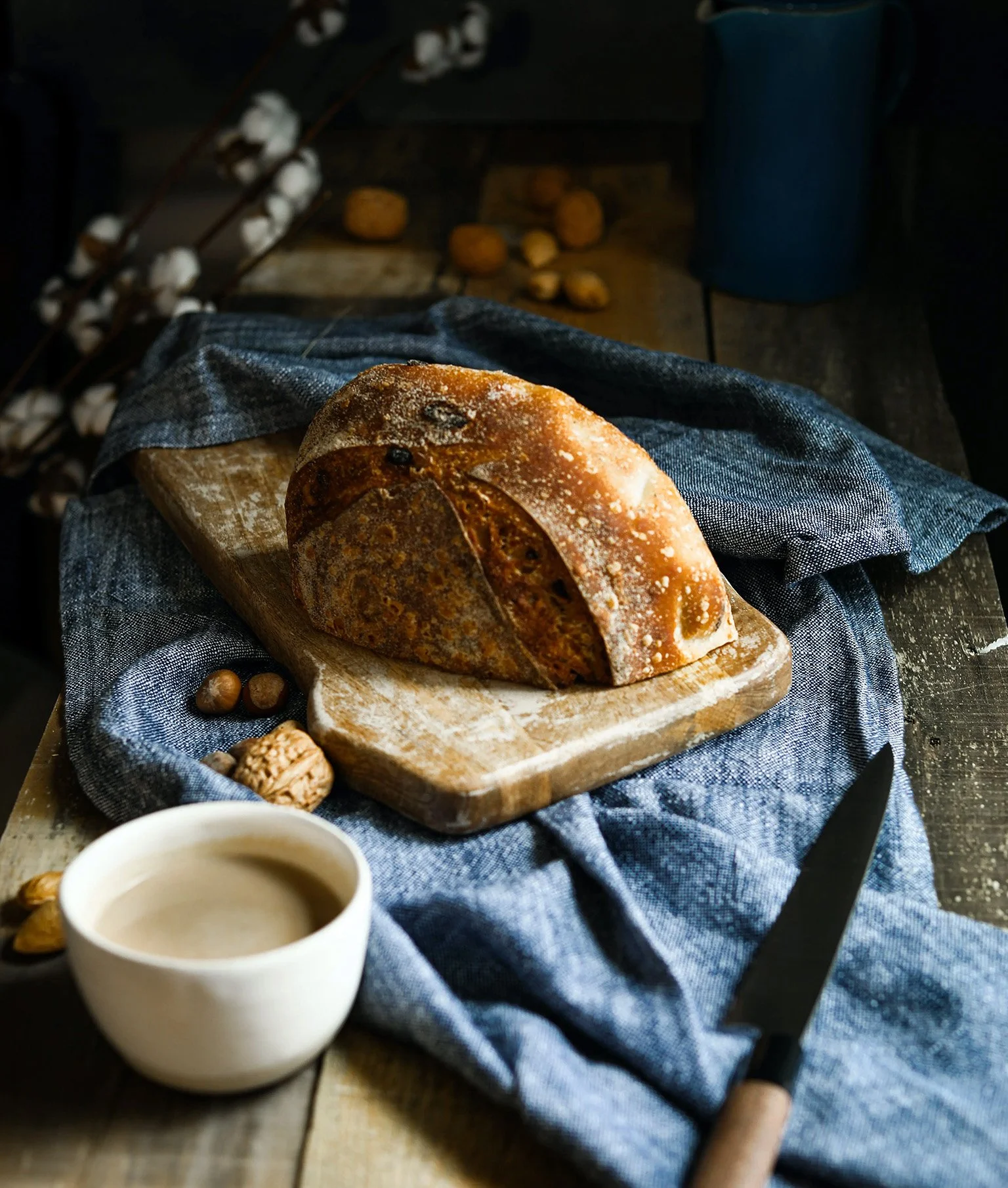 Frühstücksbrett mit angeschnittenem Brot auf blauem Tuch, ein Kaffee steht daneben.
