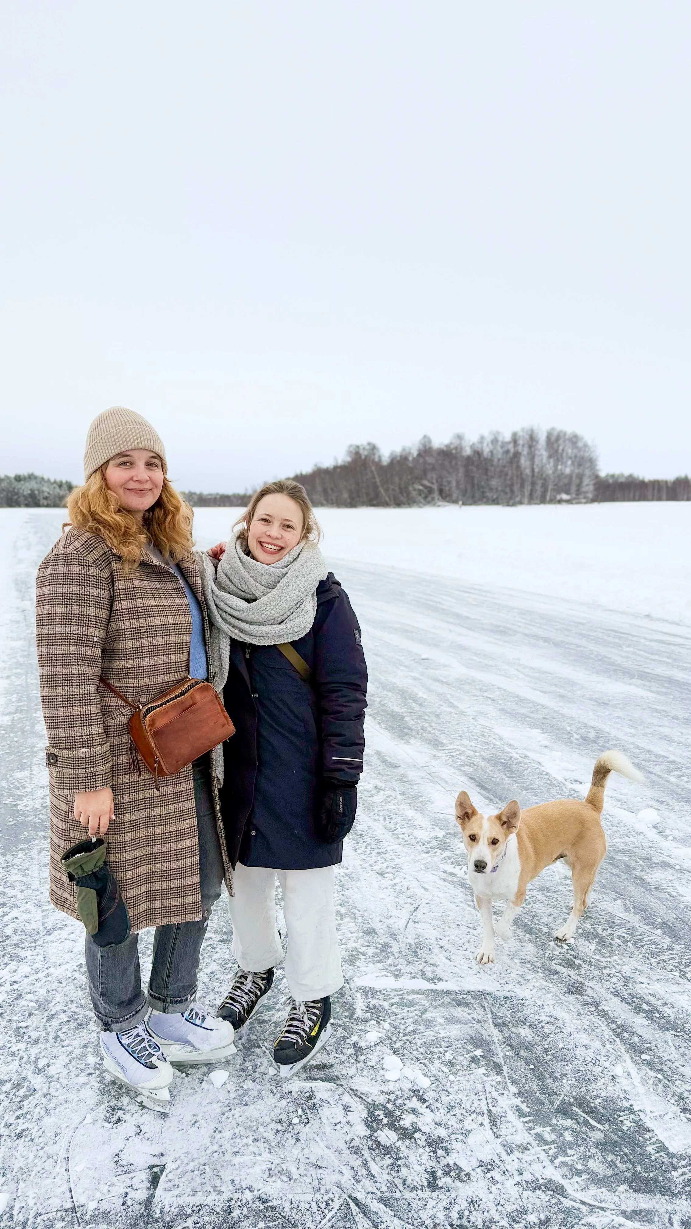 Zwei lachende Frauen, die auf einem gefrorenen See in winterlicher Kleidung stehen, mit einem Hund neben ihnen.
