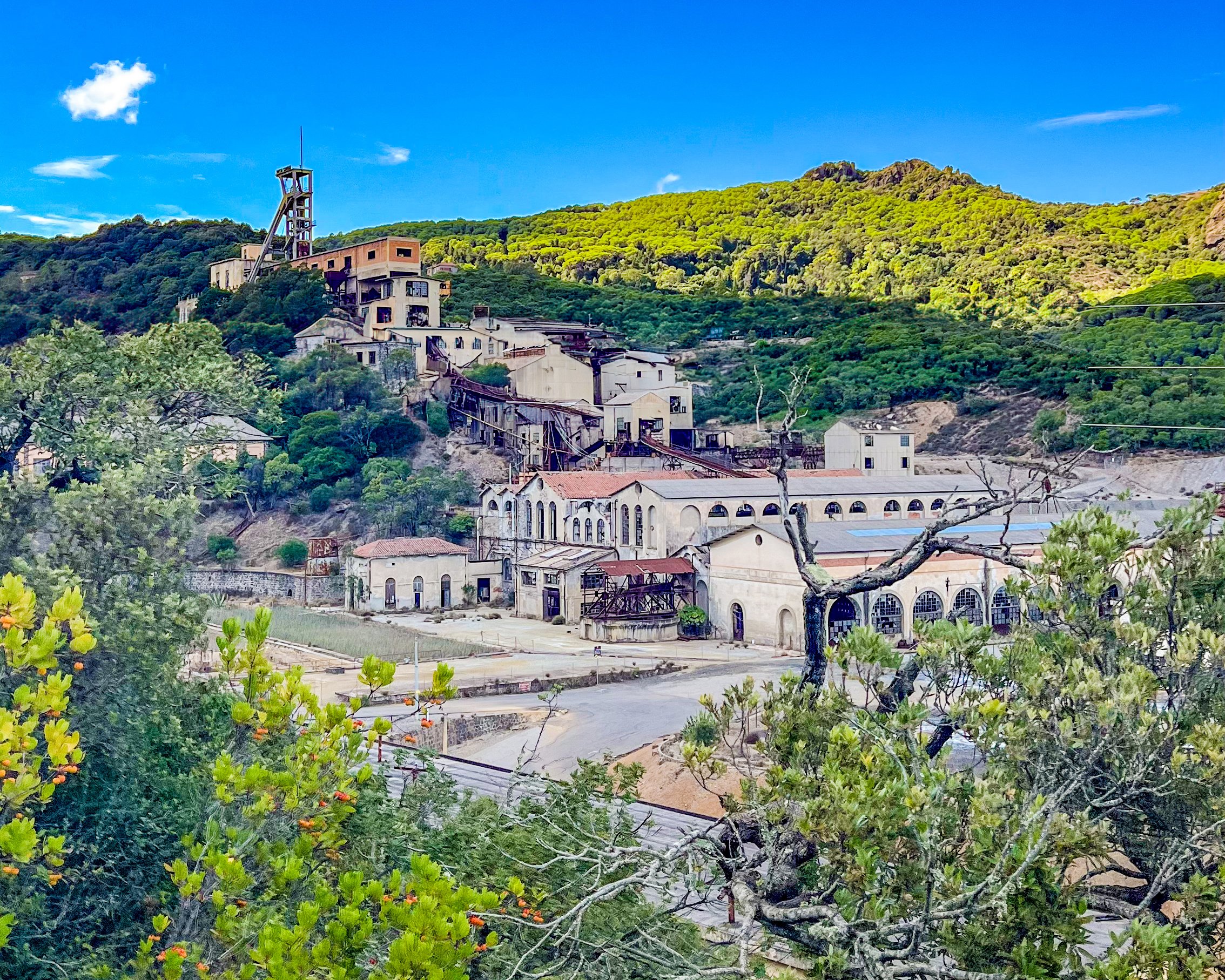 Ein Bergdorf mit alten Fabrikgebäuden und einer stillgelegten Bahn, umgeben von grünen Hügeln und Bäumen, unter einem blauen Himmel.
