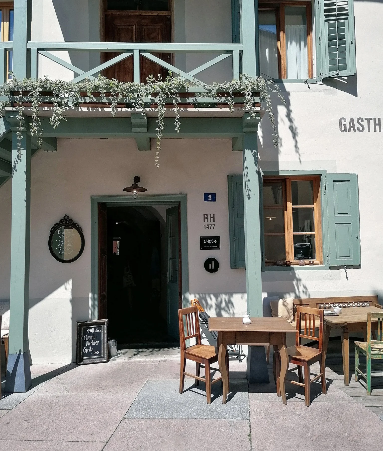 Ein Gasthaus in Lana. Grüne Fensterläden und Balkon, Holztische und Stühle davor.
