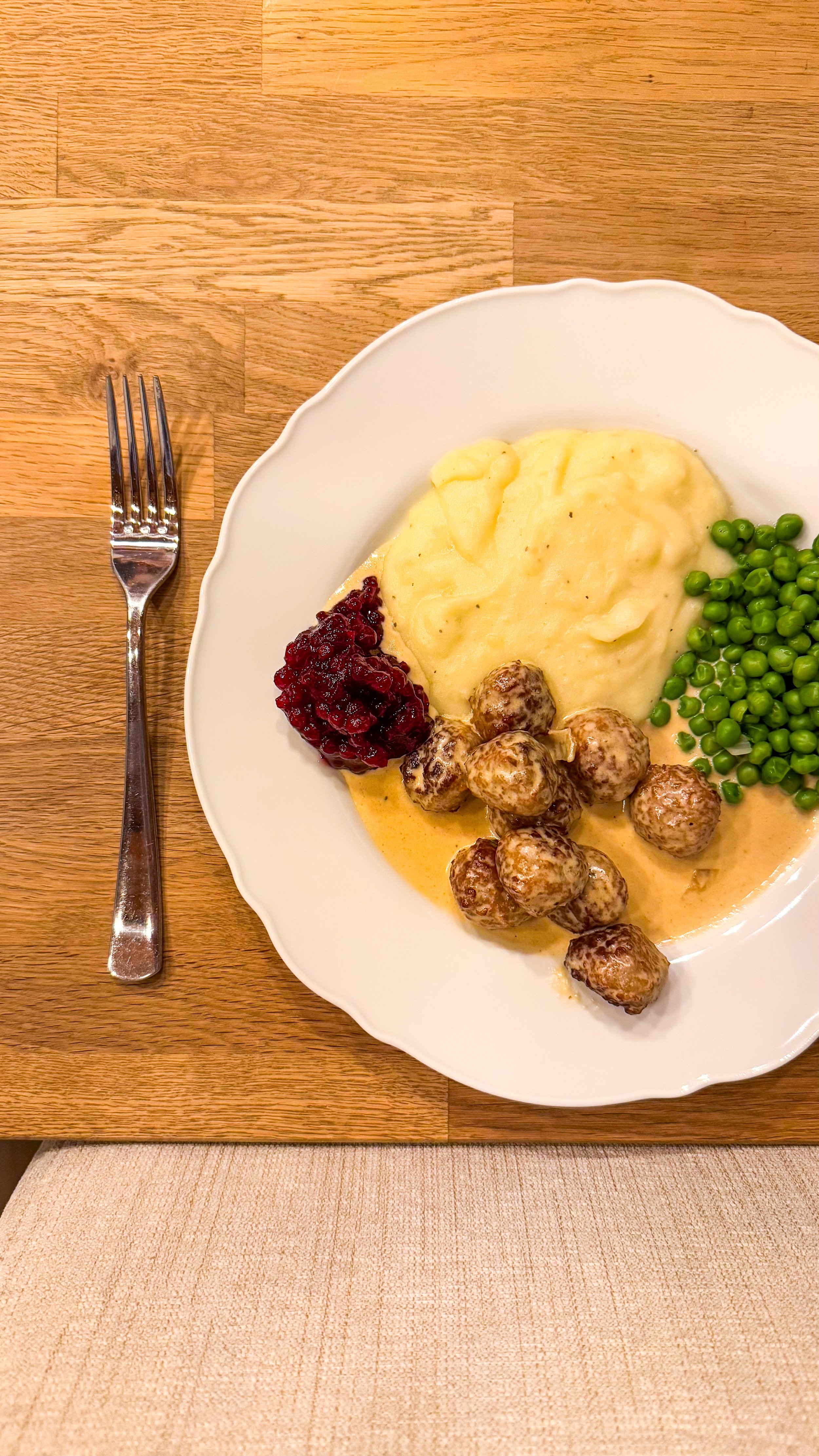 Ein Teller mit Kartoffelbrei, Preiselbeeren, Hackbällchen in Sauce und grünen Erbsen auf Holztisch.