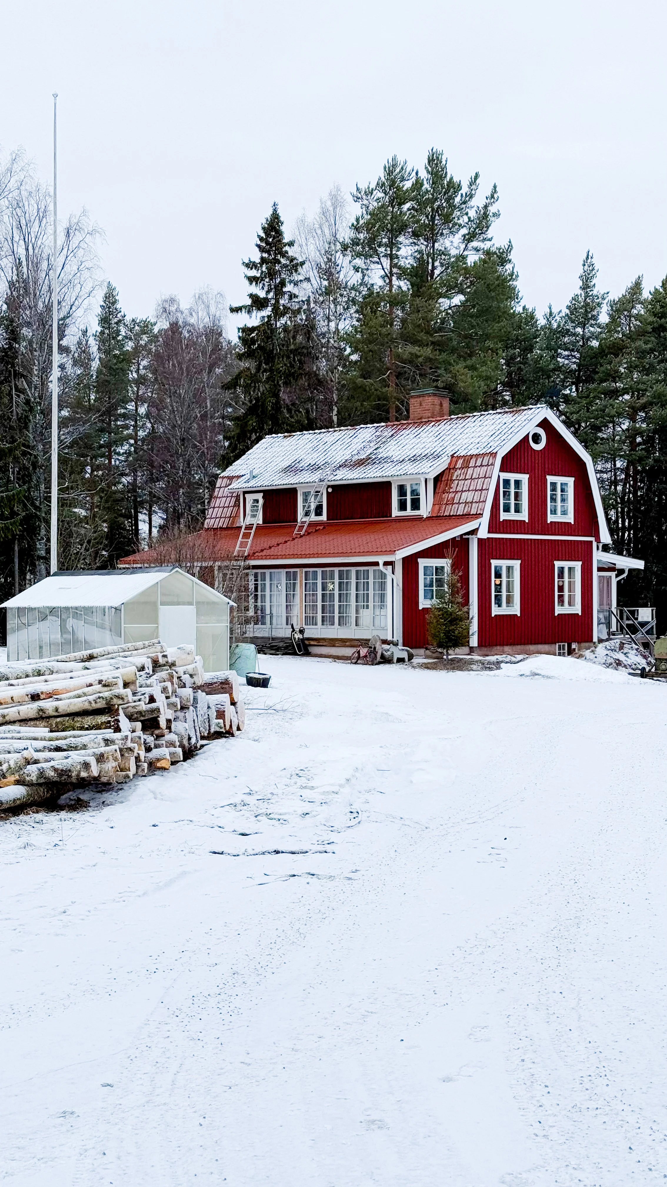 Ein rotes Holzhaus in schneebedeckter Winterlandschaft mit einem grossen Garten, einem kleinen Gewächshaus und einem Wald im Hintergrund.