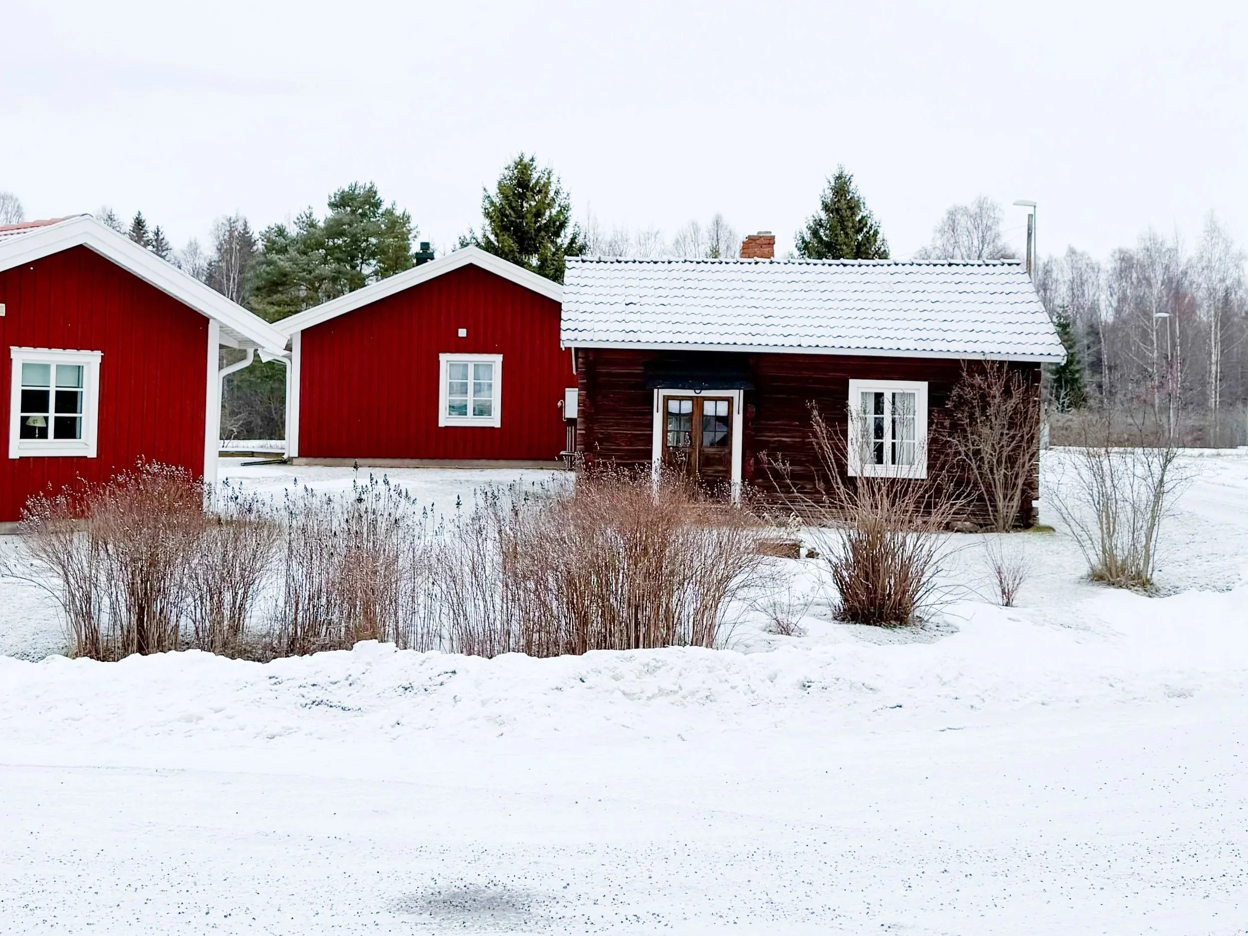 Zwei rote Holzhäuser mit weissen Fensterrahmen im Schnee, im Hintergrund Bäume, bewölkter Himmel.
