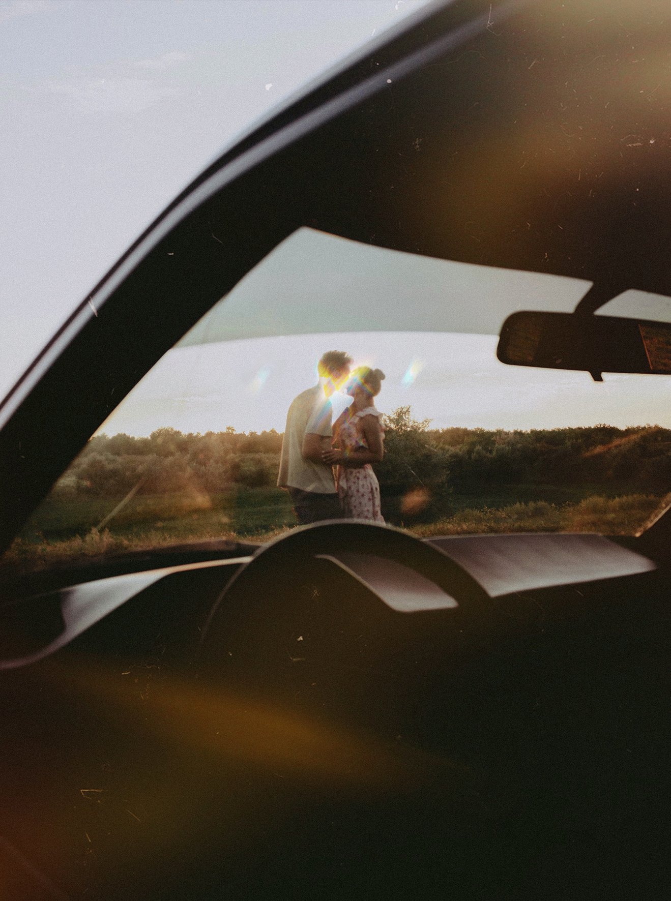 Moira und Louis küssen sich in der Abenddämmerung. Sie stehen in einem Feld, fotografiert durch die Frontscheibe ihres alten Autos.
