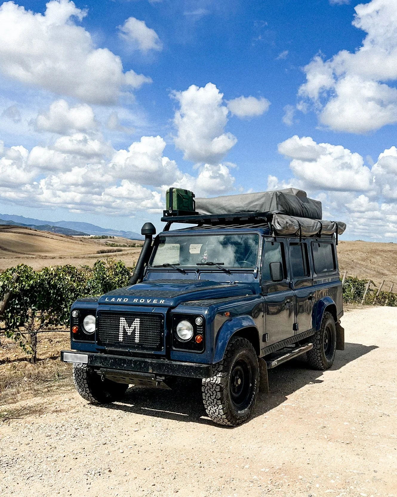 Ein blauer Land Rover Defender mit Dachzelt parkt inmitten eines sardischen Weinguts.