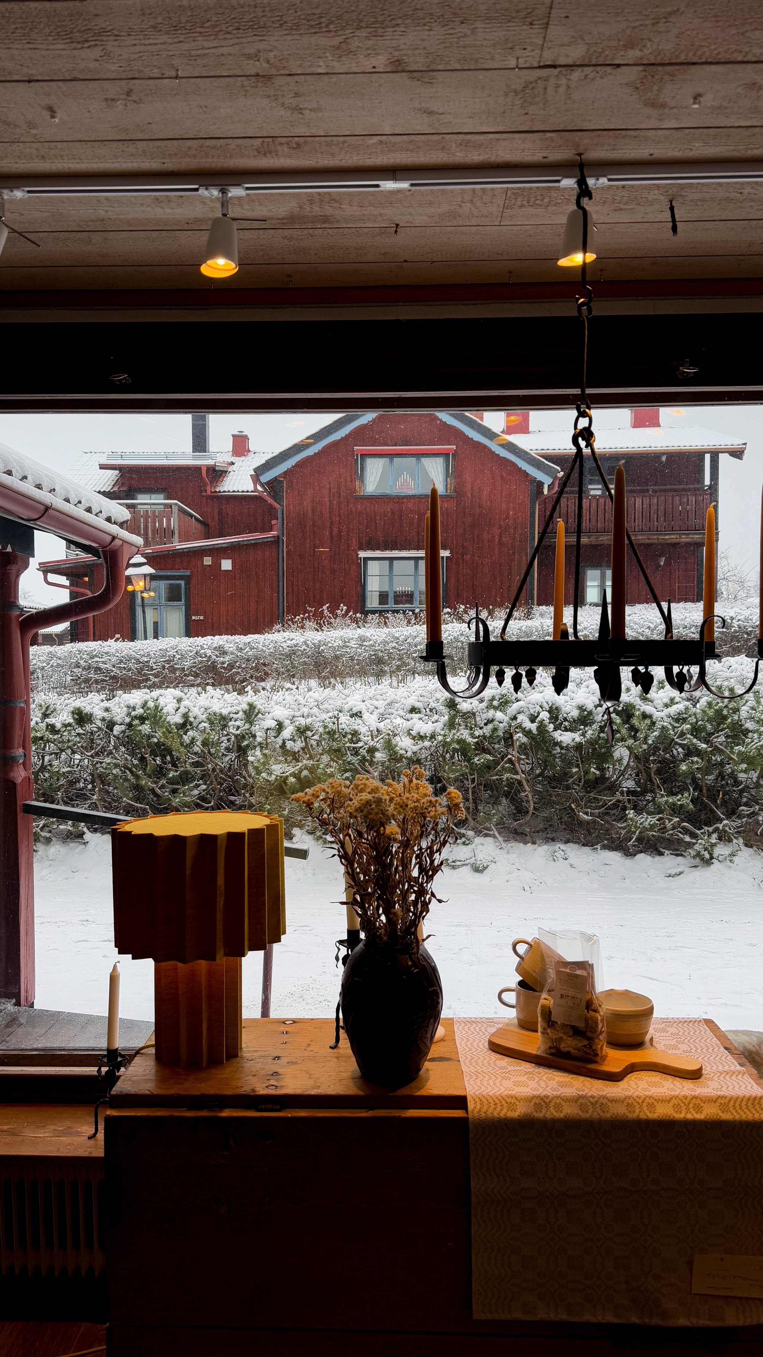 Blick aus einem Fenster auf schneebedeckte Natur mit roten Holzhäusern im Hintergrund, im Vordergrund eine Tischdekoration mit Vasen, Kerzen, einem Lampenschirm und Geschirr.