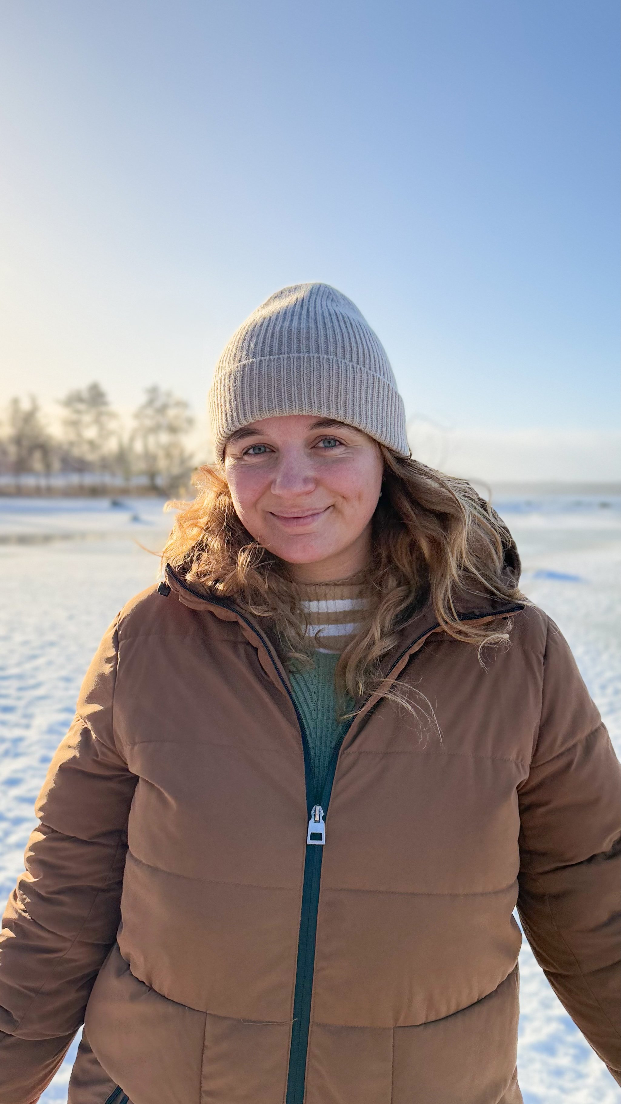 Lina trägt einen braunen Wintermantel und eine beige Wollmütze, steht im Schnee am Strand mit Blick auf das Meer bei Sonnenuntergang.