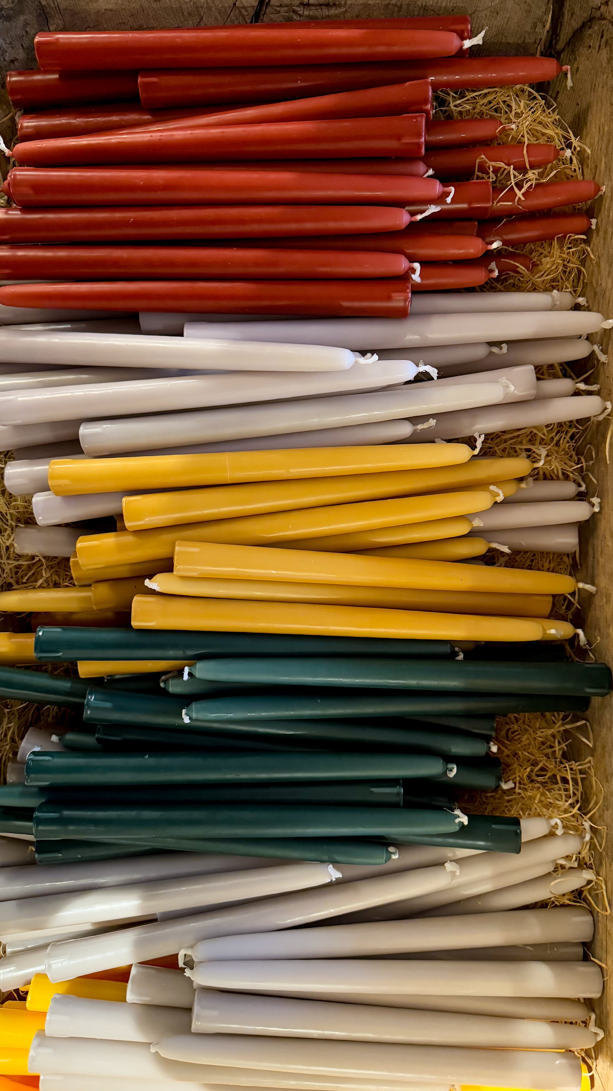 Bunte Kerzen in Rot, Weiss, Gelb, Grün, Creme, und anderen Farben, nebeneinander in einer Holzkiste mit Stroh.