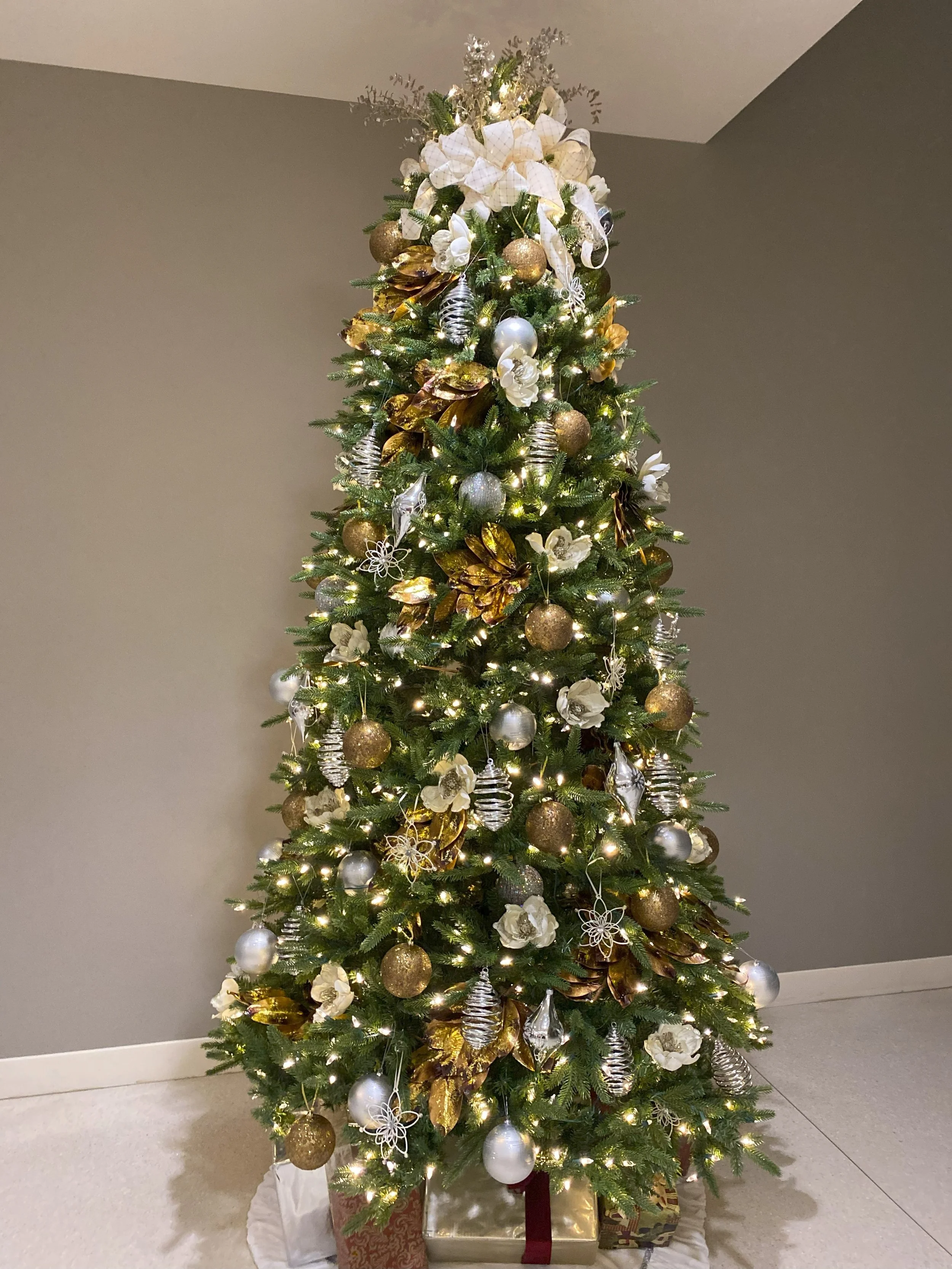 Decorated Christmas tree with gold, white, and silver ornaments, ribbons, and lights, with wrapped presents underneath.