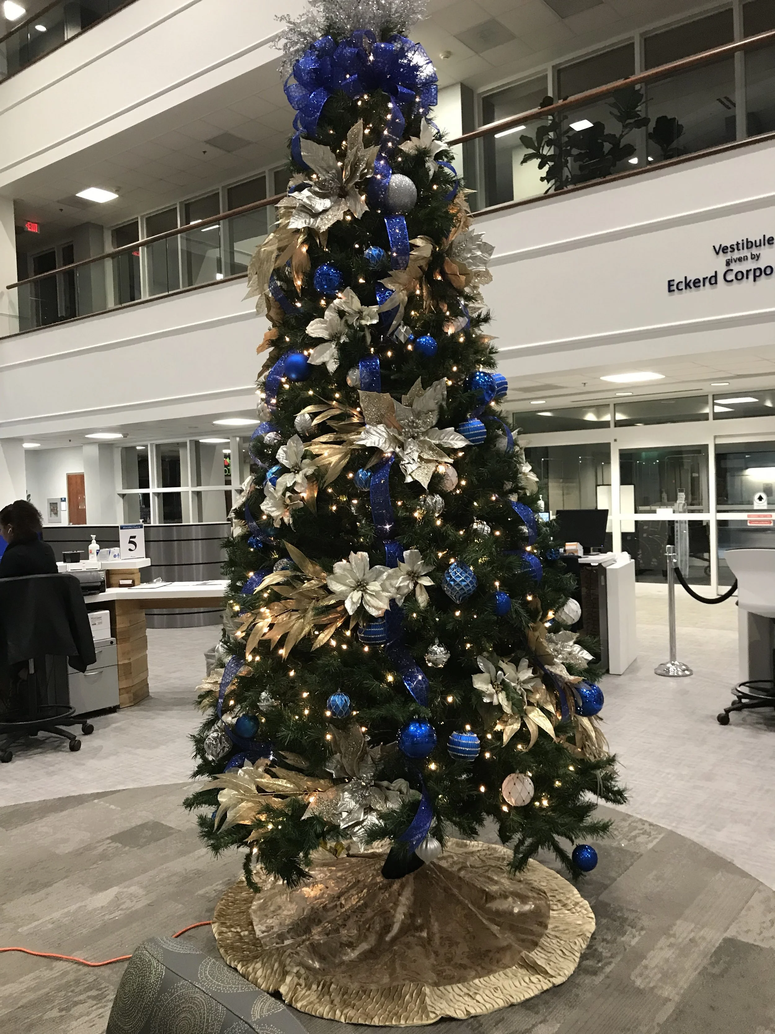 Decorated Christmas tree with blue, silver, and gold ornaments, ribbons, and lights in a modern office lobby.