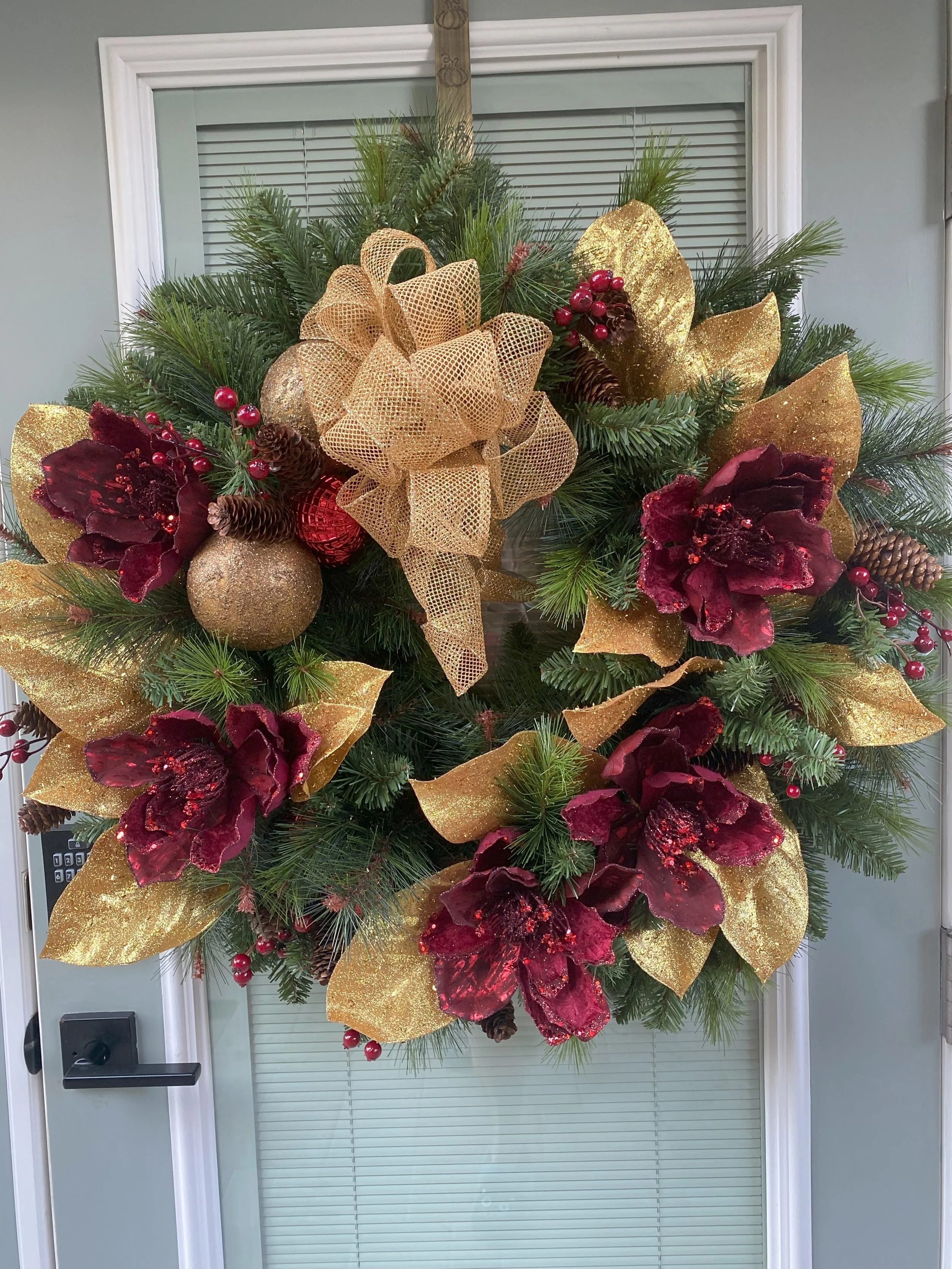Decorative Christmas wreath on a door, featuring red poinsettias, gold leaves, pinecones, red berries, and a large gold mesh bow.