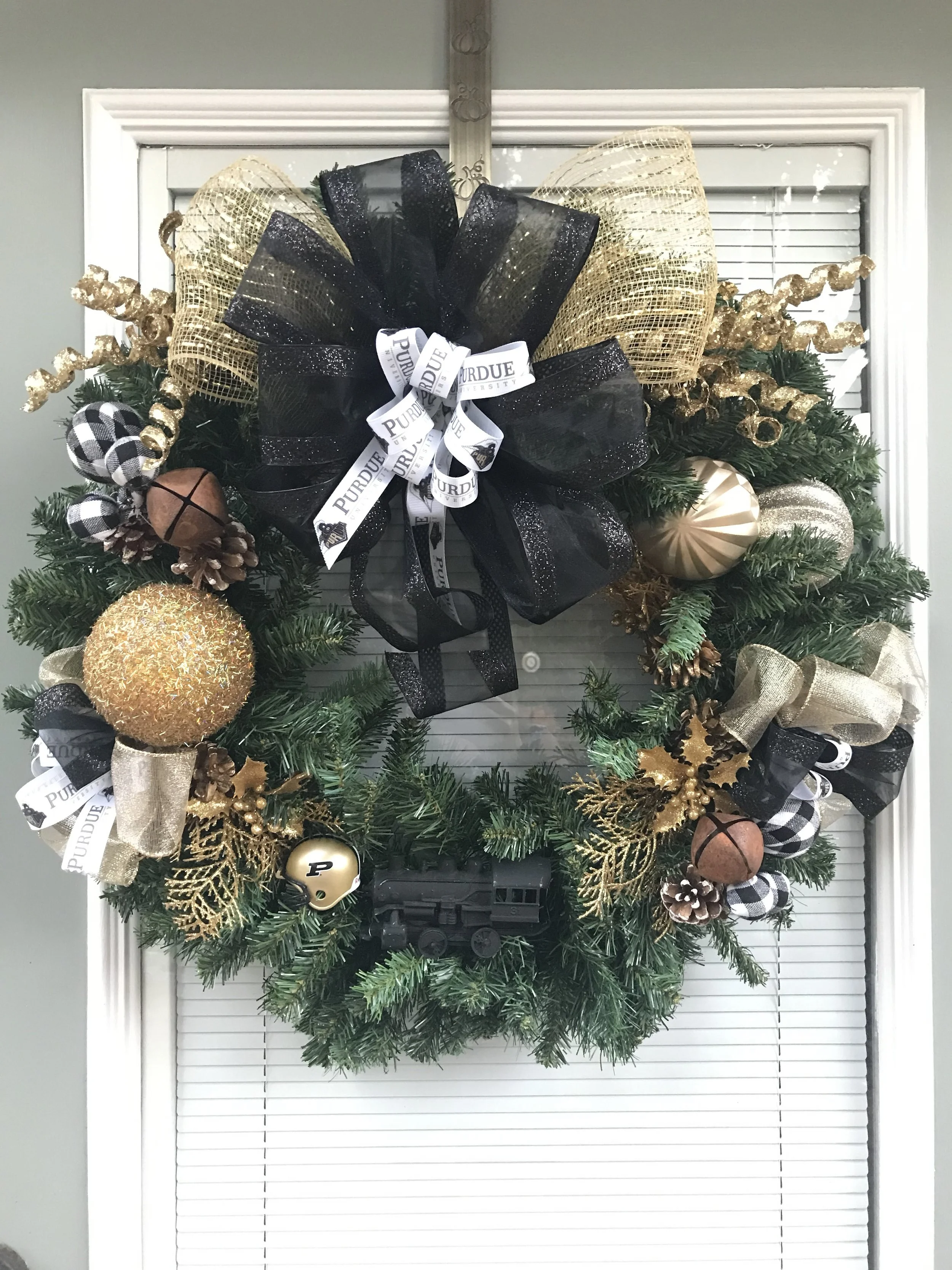 Decorated Christmas wreath hanging on a window with black, gold, and checkered ornaments, ribbons, pine cones, and a Purdue University theme, featuring a small train ornament at the bottom center.