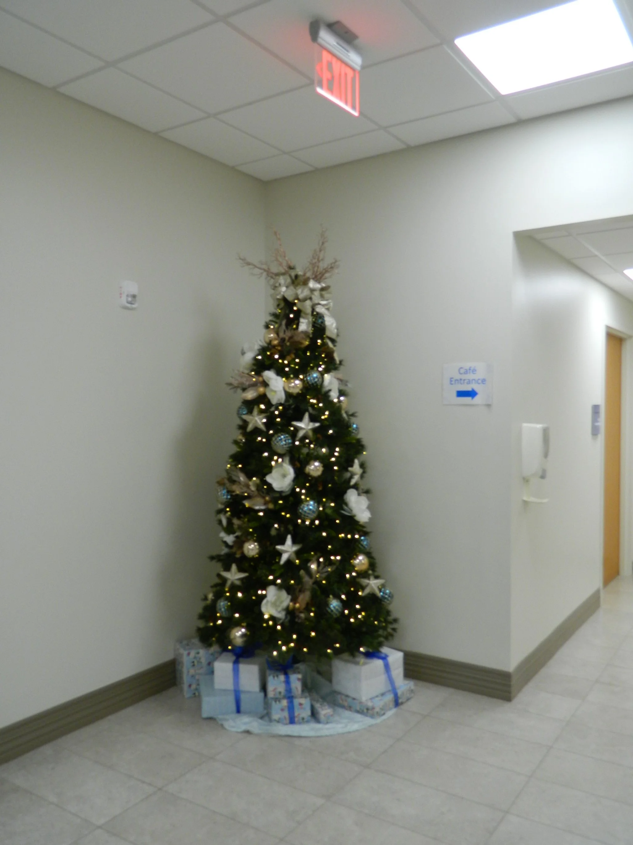 Christmas tree decorated with white and gold ornaments, lights, and a bow on top, with wrapped presents underneath, placed in a hallway near a sign for Cafe Entrance and an exit door.