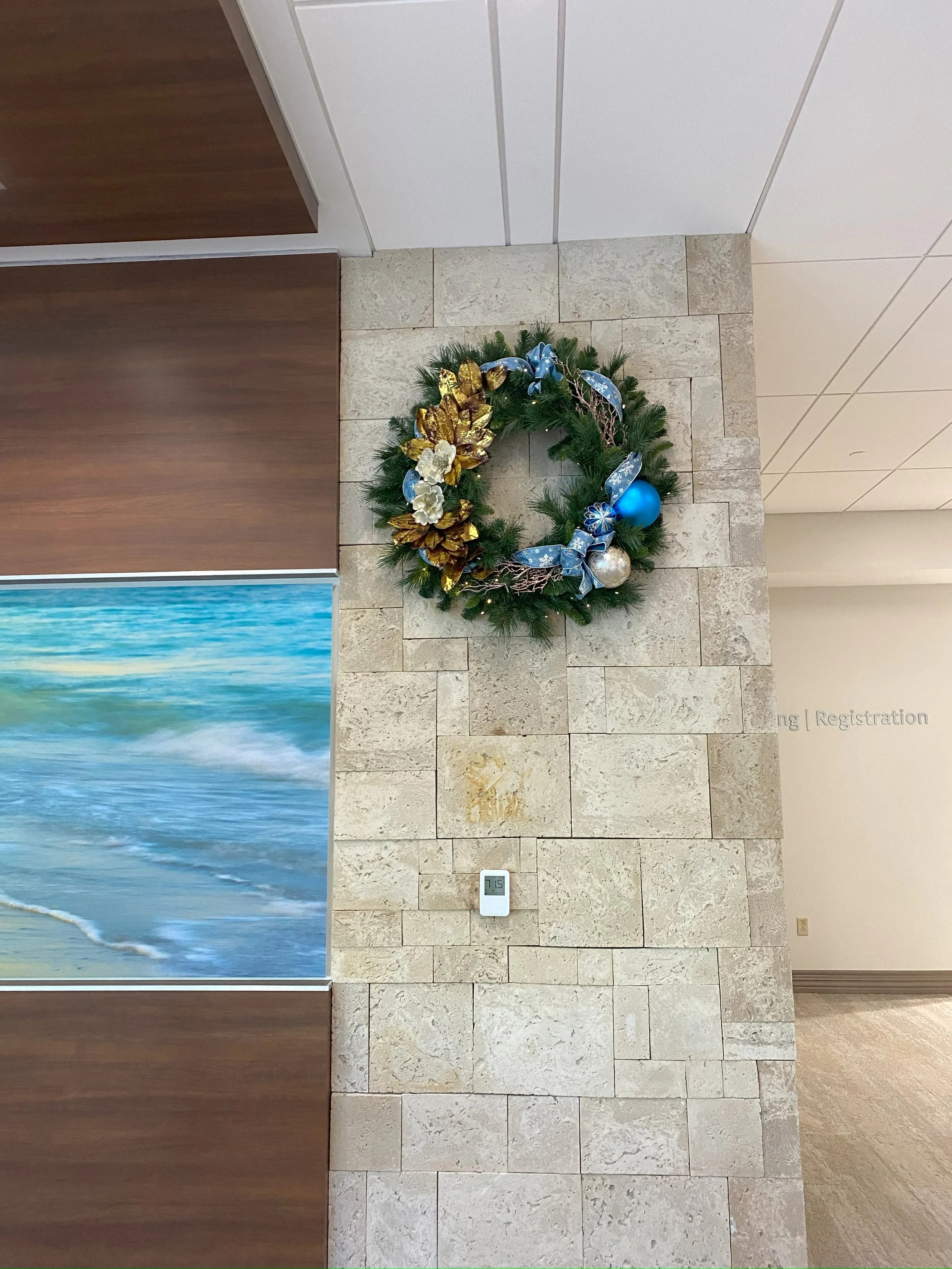 A holiday wreath decorated with gold, silver, and blue ornaments and ribbons hanging on a beige stone wall in an indoor setting.