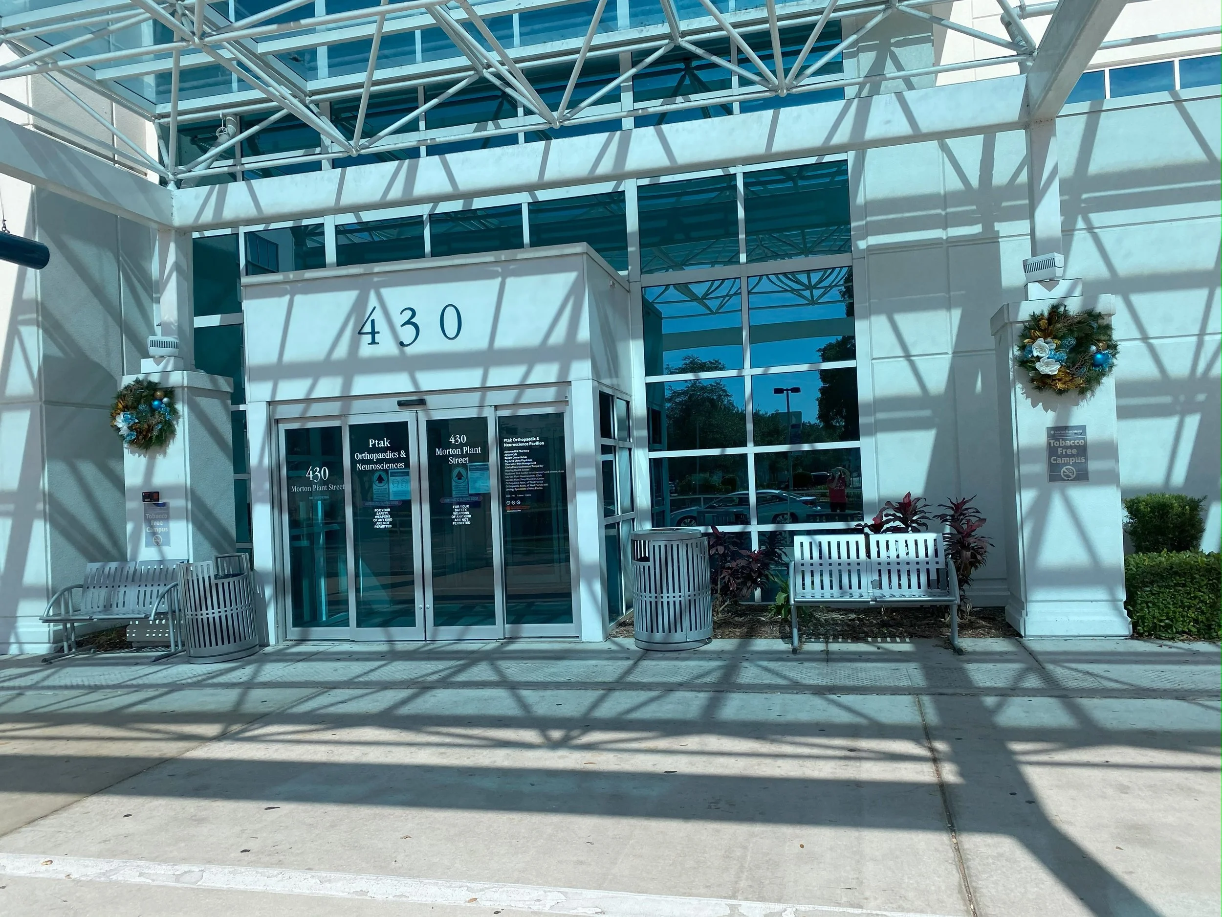 Entrance to a building with the number 430 on the front, decorated with holiday wreaths on either side, and surrounded by benches, plants, and trash cans, with clear glass windows and a metal canopy overhead.