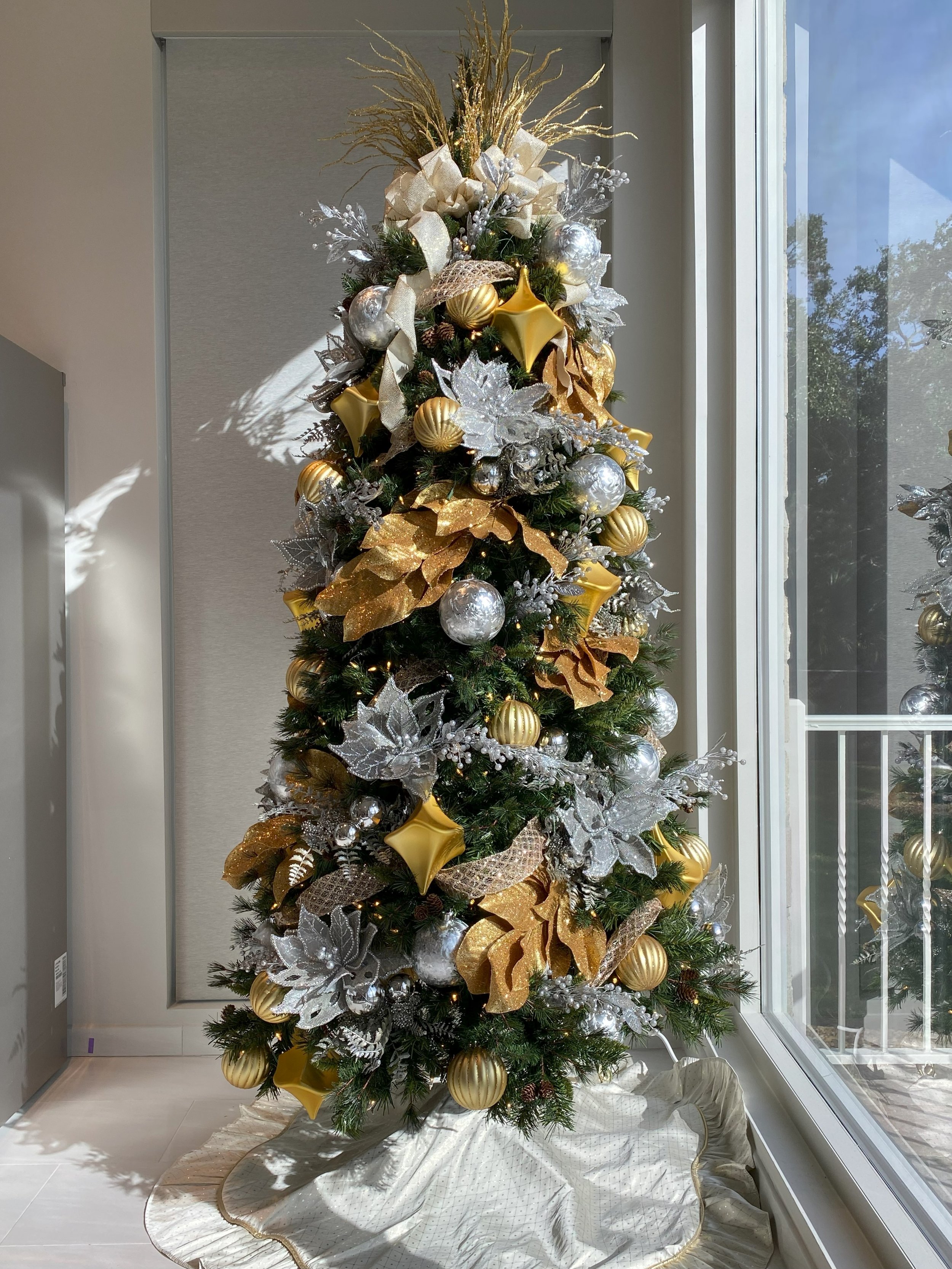 Decorated Christmas tree with gold, silver, and white ornaments, ribbons, and leaves, standing by a large window.