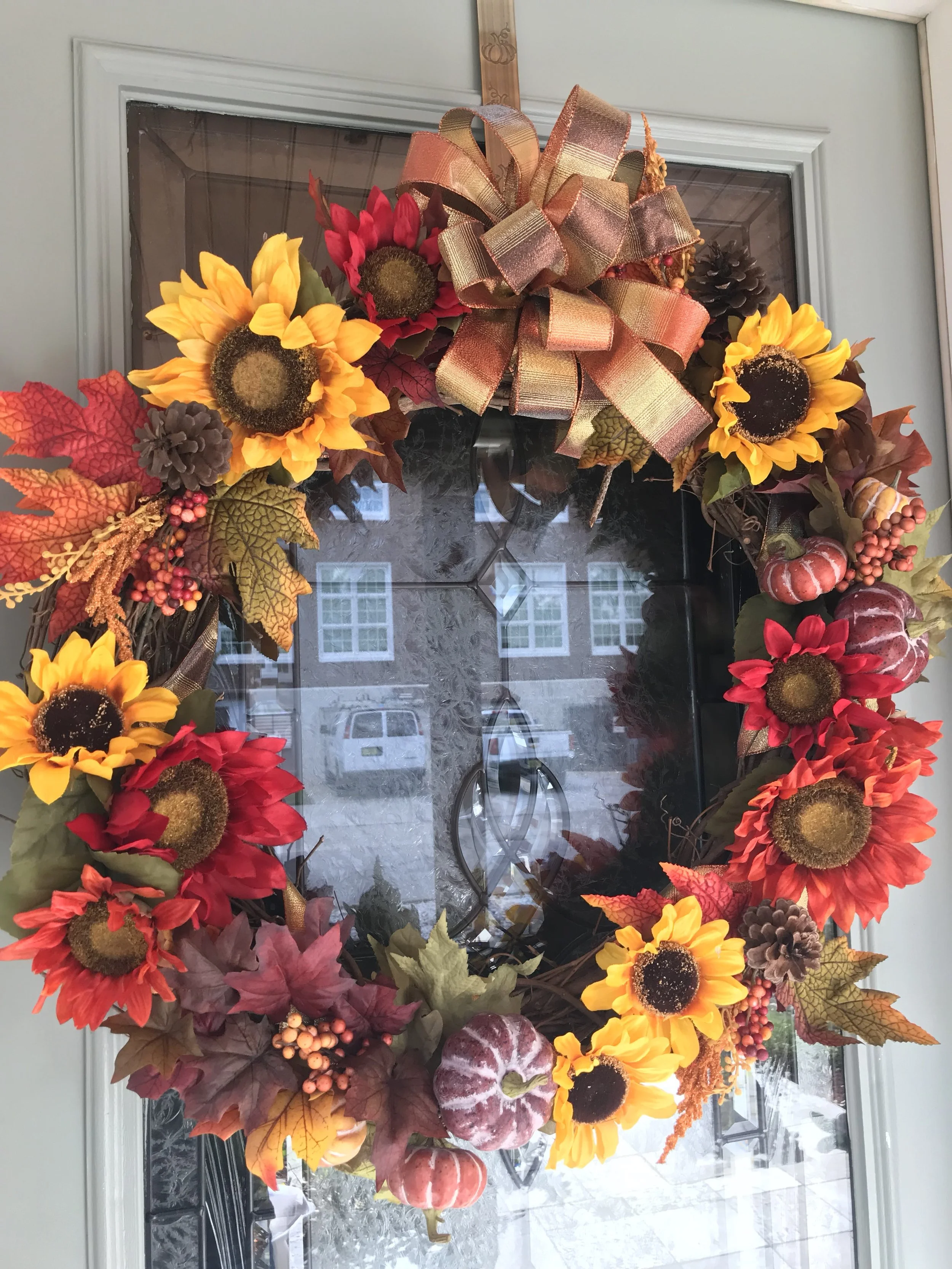Fall-themed decorative wreath with sunflowers, pumpkins, pinecones, colorful leaves, berries, and a large gold ribbon, hanging on a glass door.