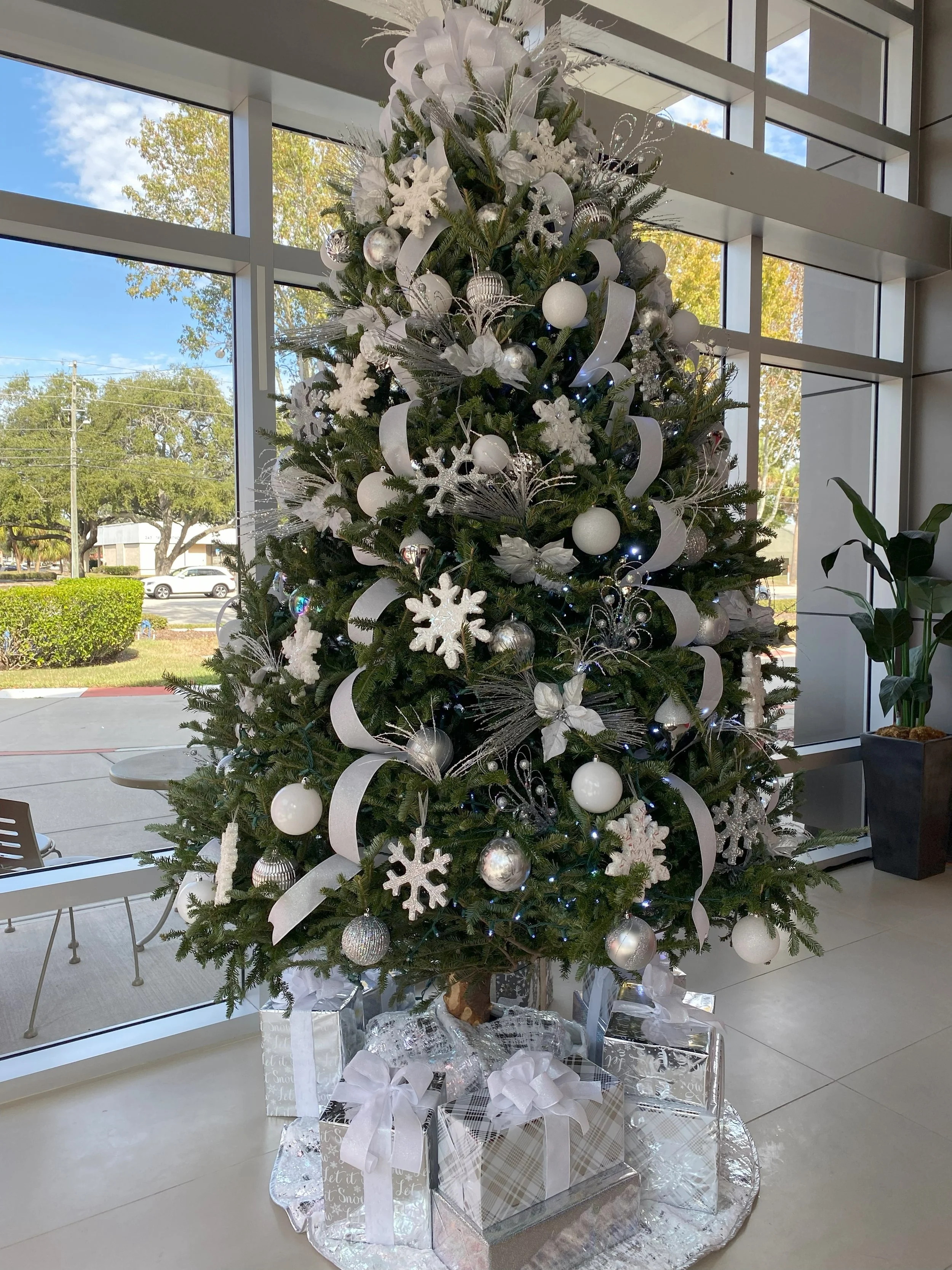 Decorated Christmas tree with white and silver ornaments, ribbons, snowflakes, and wrapped presents at the base inside a modern building with large windows.