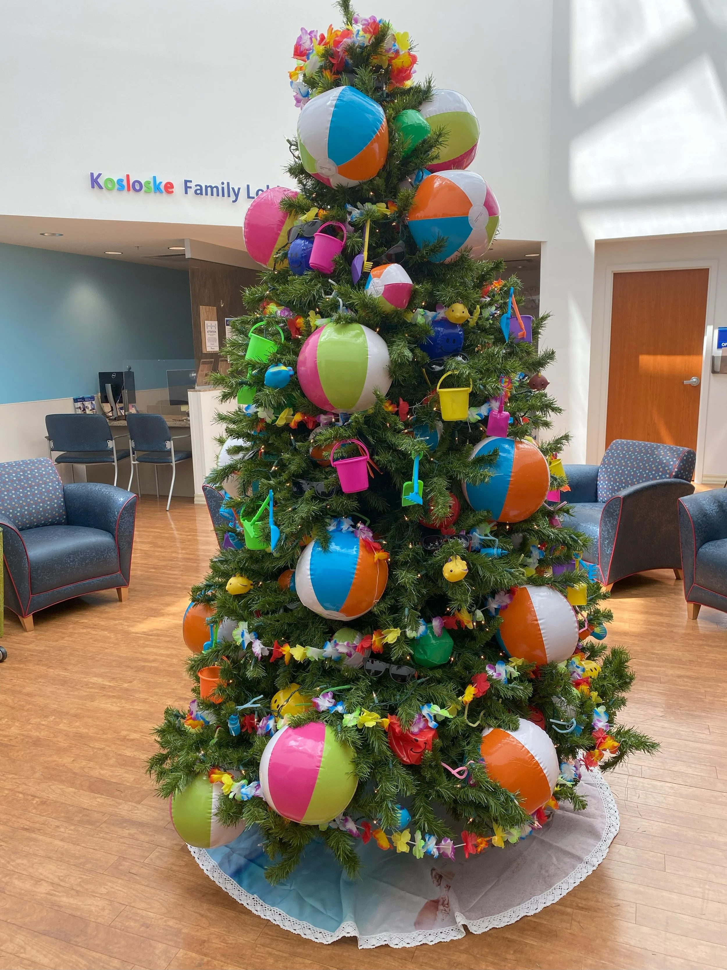 A Christmas tree decorated with colorful beach balls, small watering cans, sunglasses, and inflatable toys, placed in a lobby area with chairs and a sign that reads 'Kosloske Family Lo' in the background.