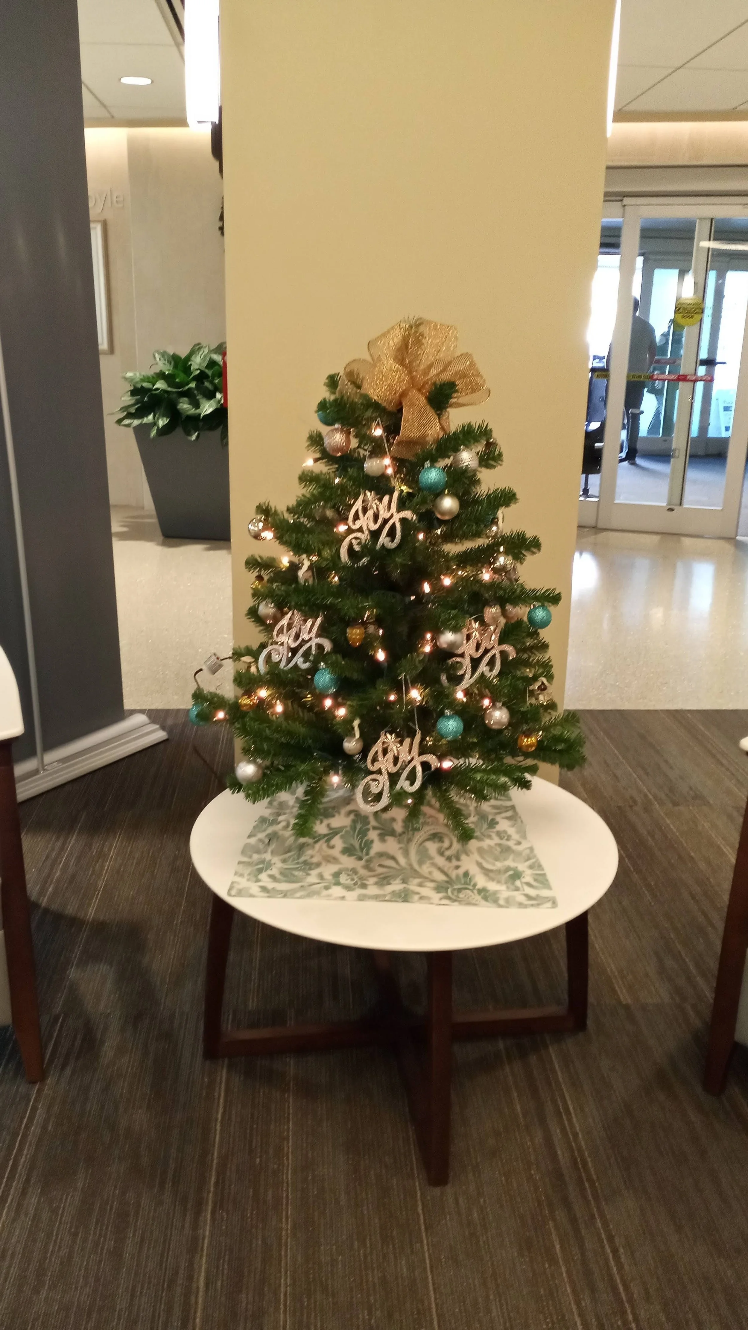 Small decorated Christmas tree with gold bow on top, ornaments, and lights, placed on a white table in a public indoor space.