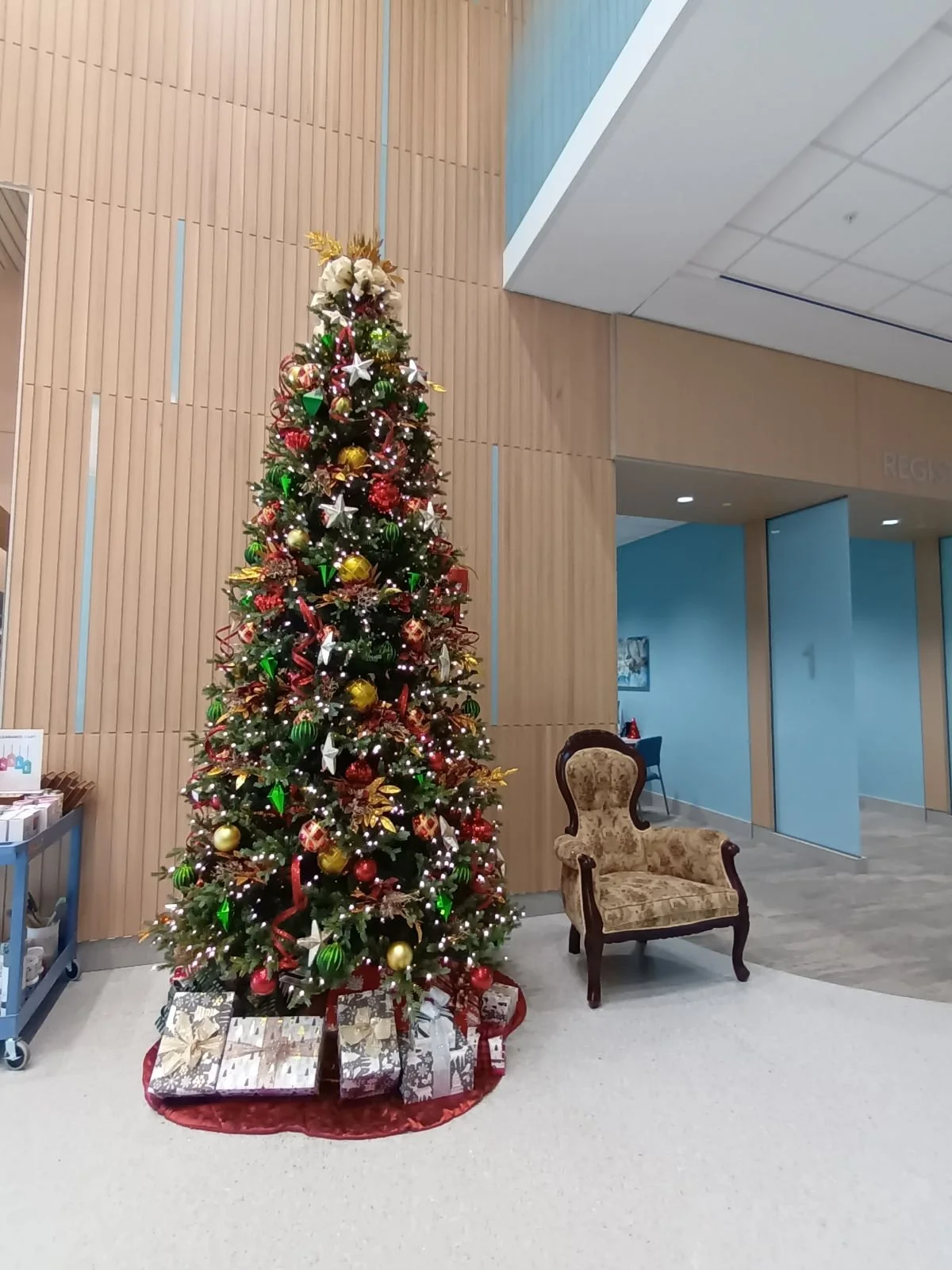Decorated Christmas tree with presents underneath, situated in a modern indoor space with a wooden wall and a vintage armchair nearby.