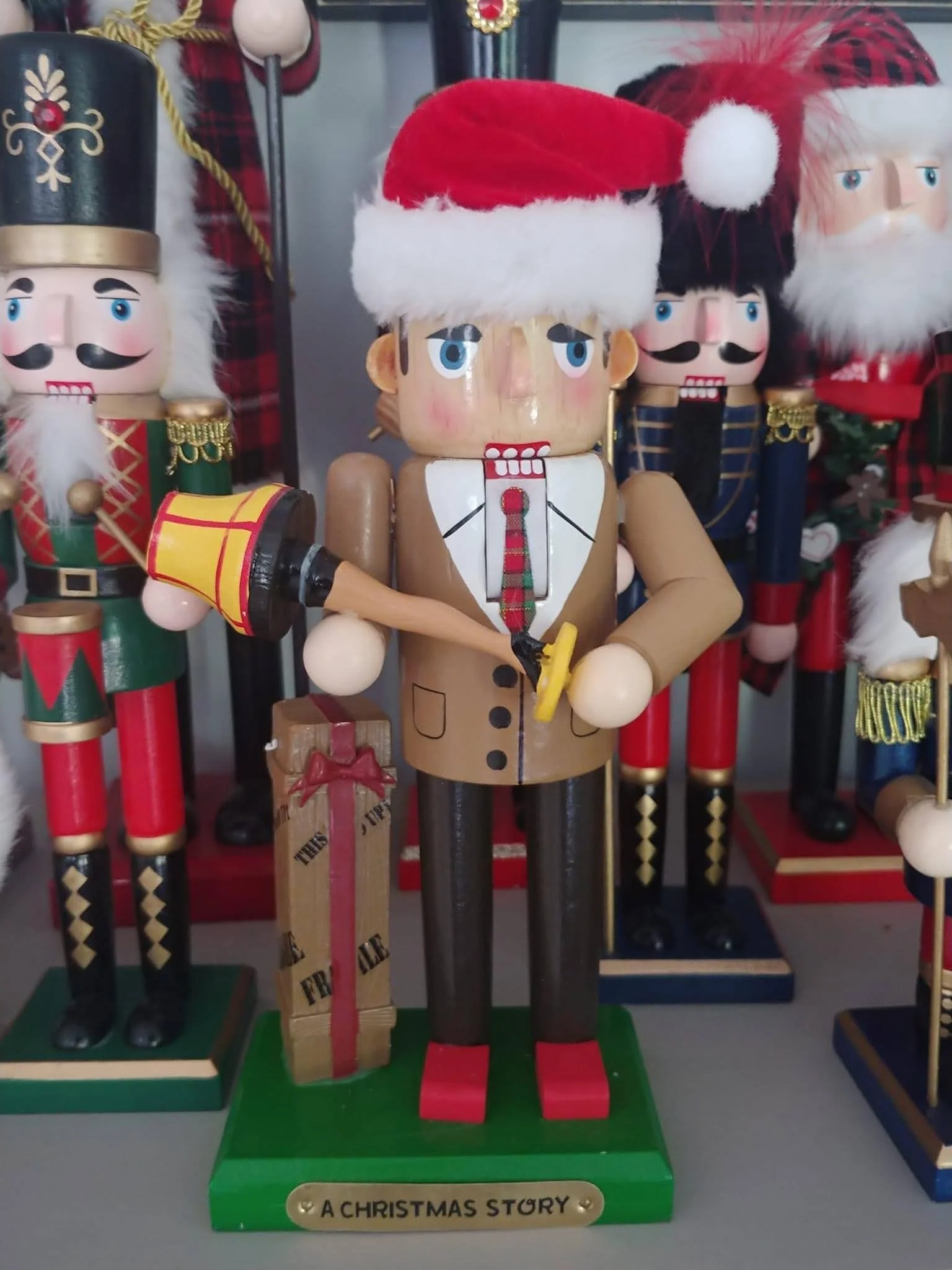 Wooden nutcracker figurine wearing a Santa hat, holding a yellow axe, with a green base labeled "A Christmas Story," surrounded by other nutcracker figurines dressed in Christmas attire.