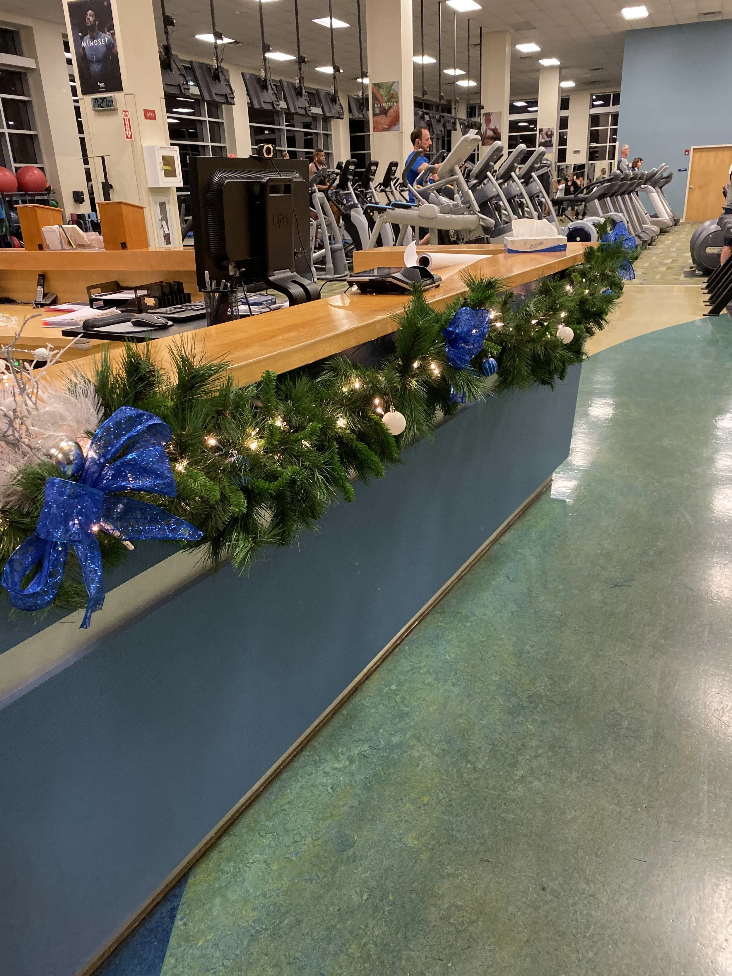 Indoor gym reception area decorated with Christmas garland, blue ribbons, and ornaments. Reception desk with a computer, telephone, and various items. Workout equipment in the background with people using treadmills and ellipticals.